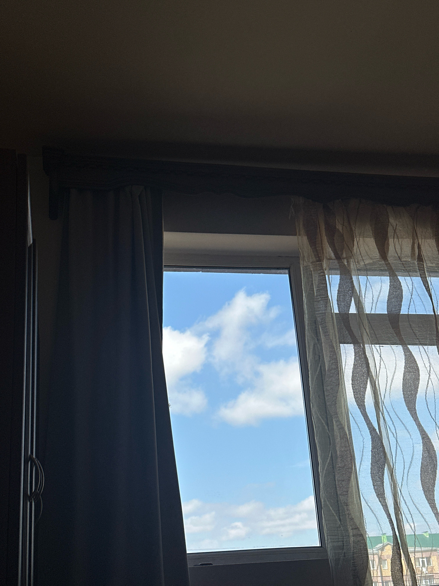 The image depicts an interior view looking out a window. On the left, a dark, opaque curtain is drawn partially across the window, obscuring a portion of the view. To the right of the dark curtain, a sheer, patterned curtain with a wavy design is visible, allowing light to pass through. Through the windows, a bright blue sky with scattered white clouds is visible. In the lower portion of the frame, buildings with green and brown rooftops can be seen, suggesting an urban environment. The perspective suggests the viewpoint is from a residential or commercial building, likely on an upper floor. The scene appears to be captured during daylight. The specific location in Gyumri, Armenia cannot be determined from the image alone.