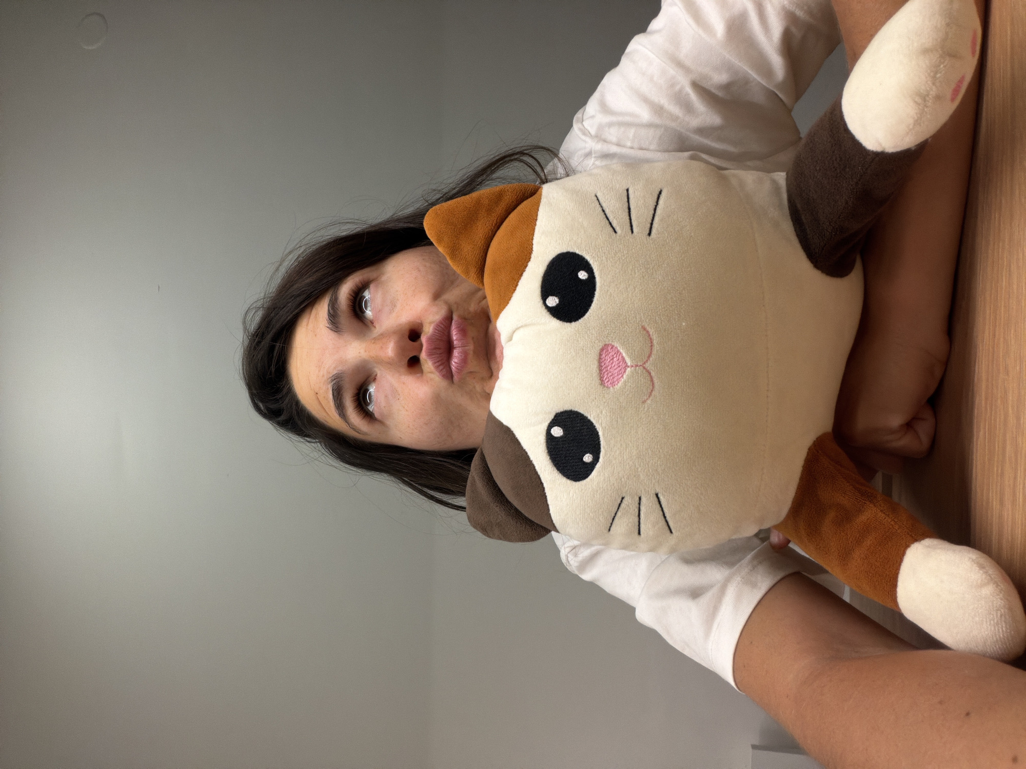 A woman with dark hair is holding a plush cat toy. She has fair skin and is wearing a white t-shirt. The cat toy is beige with brown ears and paws, a pink nose, black eyes, and black whiskers. The background is a gray wall on the left and a light brown wall on the right. No information about the location in the city of Budva, Montenegro is discernible.