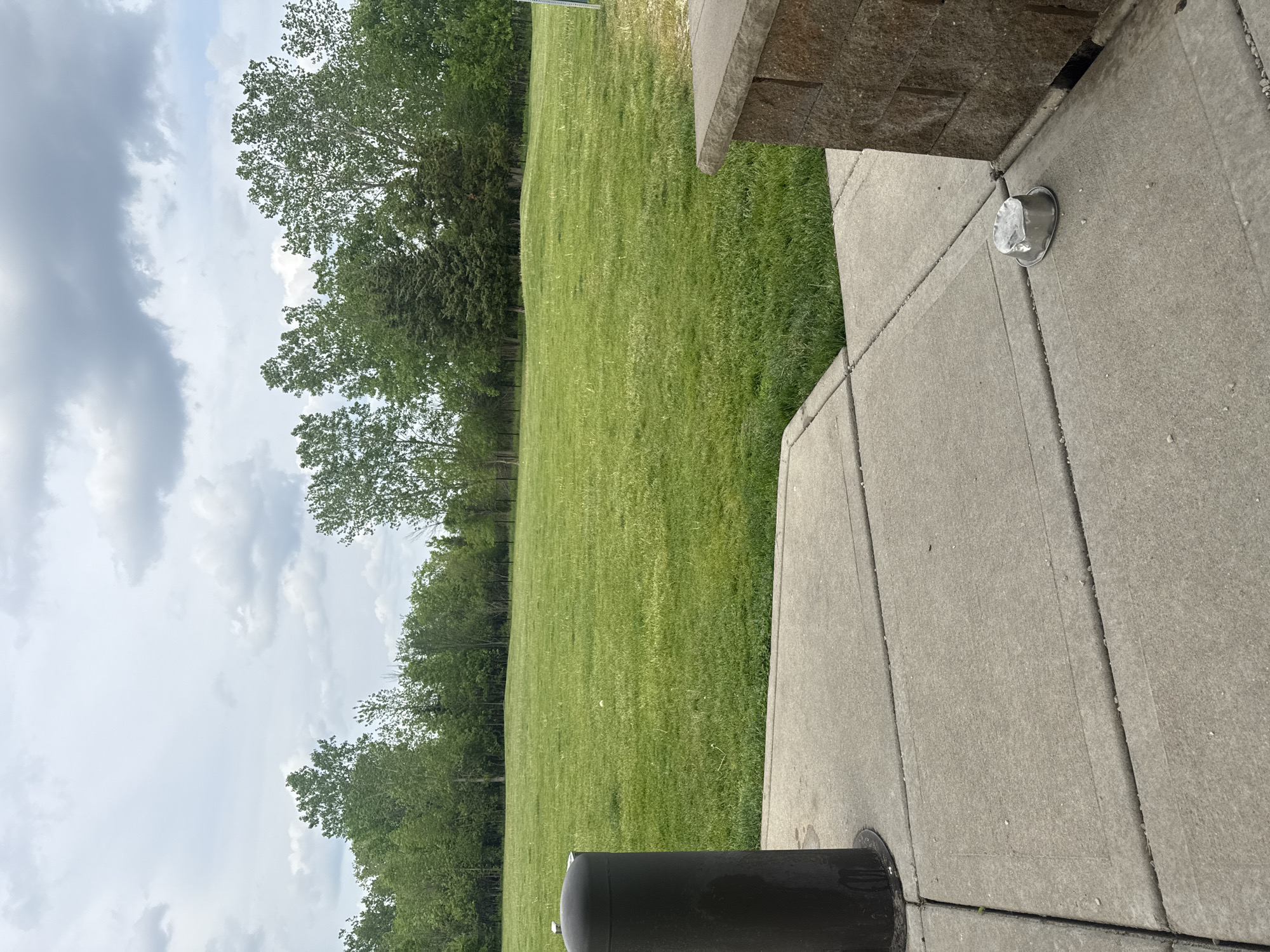 The image shows a grassy field next to a paved area, likely a sidewalk or patio. The field is green and well-maintained, with a line of trees along the edge. The paved area is made of concrete slabs with a small metal fixture set into the concrete. A cylindrical object, possibly a trash can or other type of receptacle, is partially visible in the foreground. The sky is cloudy and overcast.
The scene appears to be in Glenview, United States of America, based on the geographical information provided.