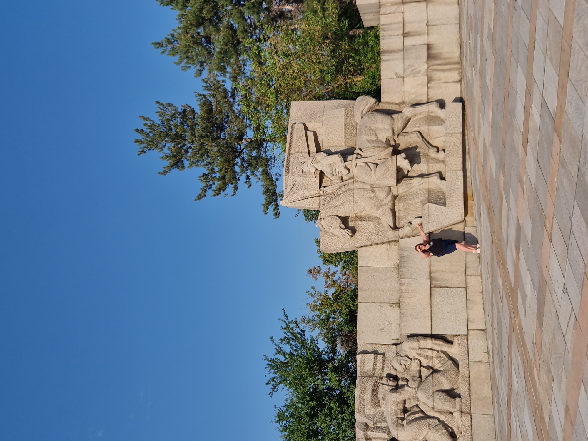 A woman in a dark blue dress and white shoes stands to the left of a large stone sculpture. The sculpture depicts figures on horseback and other figures in relief. The woman is posing for a picture, with her left hand resting on her hip and her right arm extended towards the sculpture. The background consists of a clear blue sky and several green trees. The scene is set in Panagyurishte, Bulgaria, in front of the Panagyurishte Memorial. The ground is paved with large, square tiles.