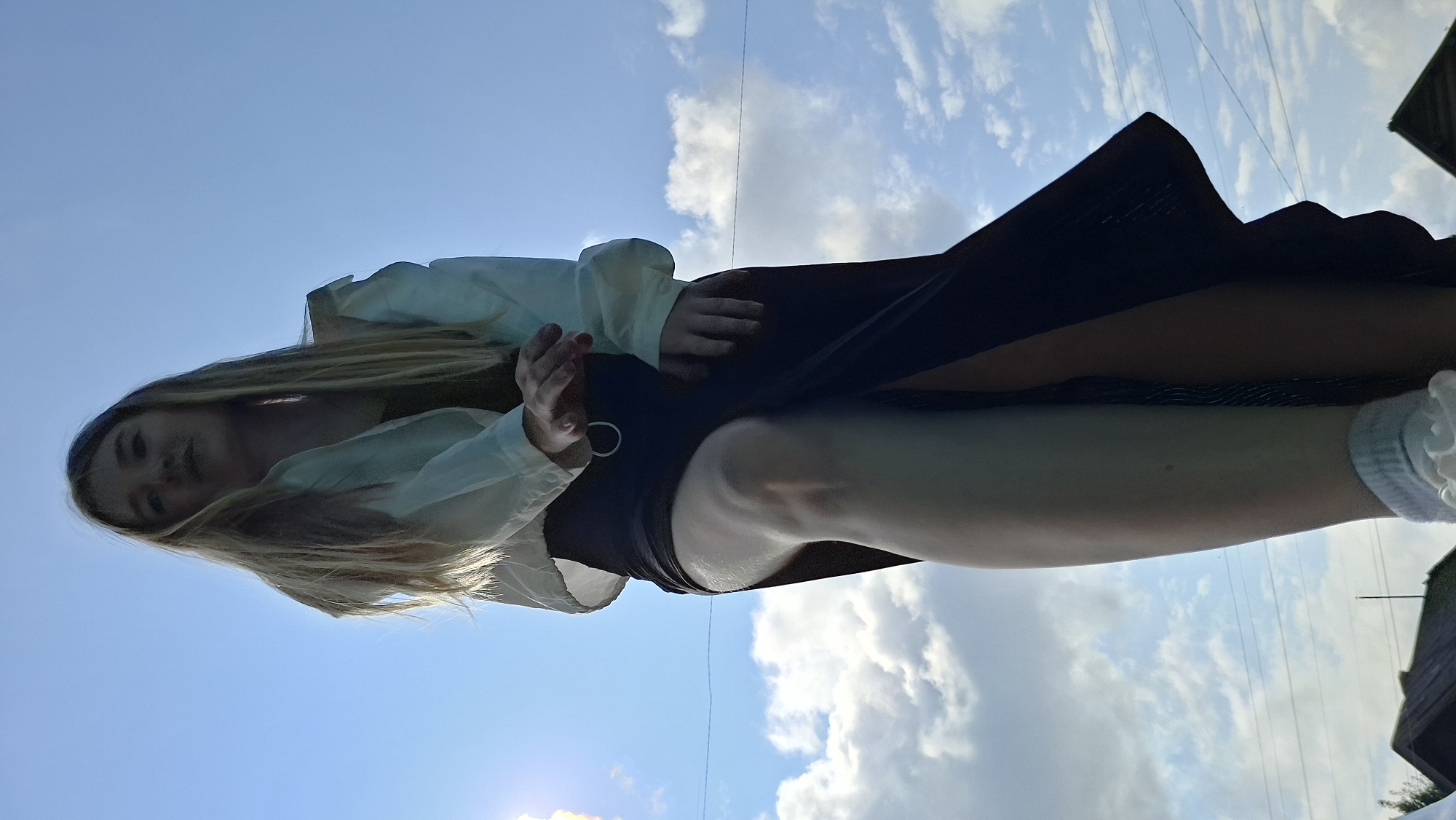 A young woman with long blonde hair is standing against a bright blue sky with scattered clouds. She is wearing a light-colored jacket over a darker top and a long, dark skirt with a high slit. Her left leg is bent, with her knee prominently visible. She has on white socks with ruffled edges. In the background, the tops of two dark-colored roofs are visible, suggesting an outdoor setting. There are also thin dark lines in the sky, possibly power lines. The image is taken from a low angle, looking up at the woman. The location is identified as Bolshoy Bukor, Russia.