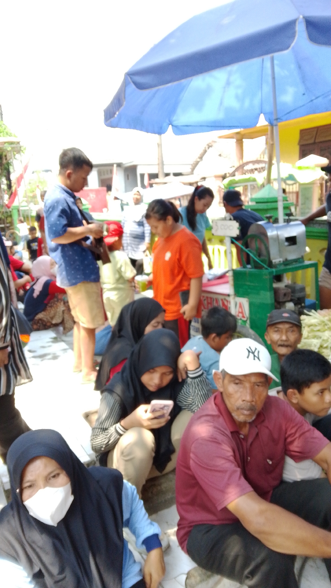In Mancar, Indonesia, under a bright sky, a group of people are gathered outdoors, likely at a market or public event. A large blue tarp or umbrella provides partial shade. In the foreground, a woman wearing a black hijab and a white face mask looks towards the camera. To her right, a man in a maroon collared shirt and dark pants sits facing forward. Behind them, several other individuals are visible. A woman in a black hijab is looking at her phone, and next to her, a person in a light blue shirt is also engaged with their device. A man in an orange shirt stands with his back slightly turned. Further back, a man in a blue patterned shirt appears to be interacting with something in his hands, possibly a camera. A green sugarcane juicing machine is visible in the background, suggesting the sale of fresh juice. The overall scene depicts a lively, casual gathering of people in an outdoor Indonesian setting.