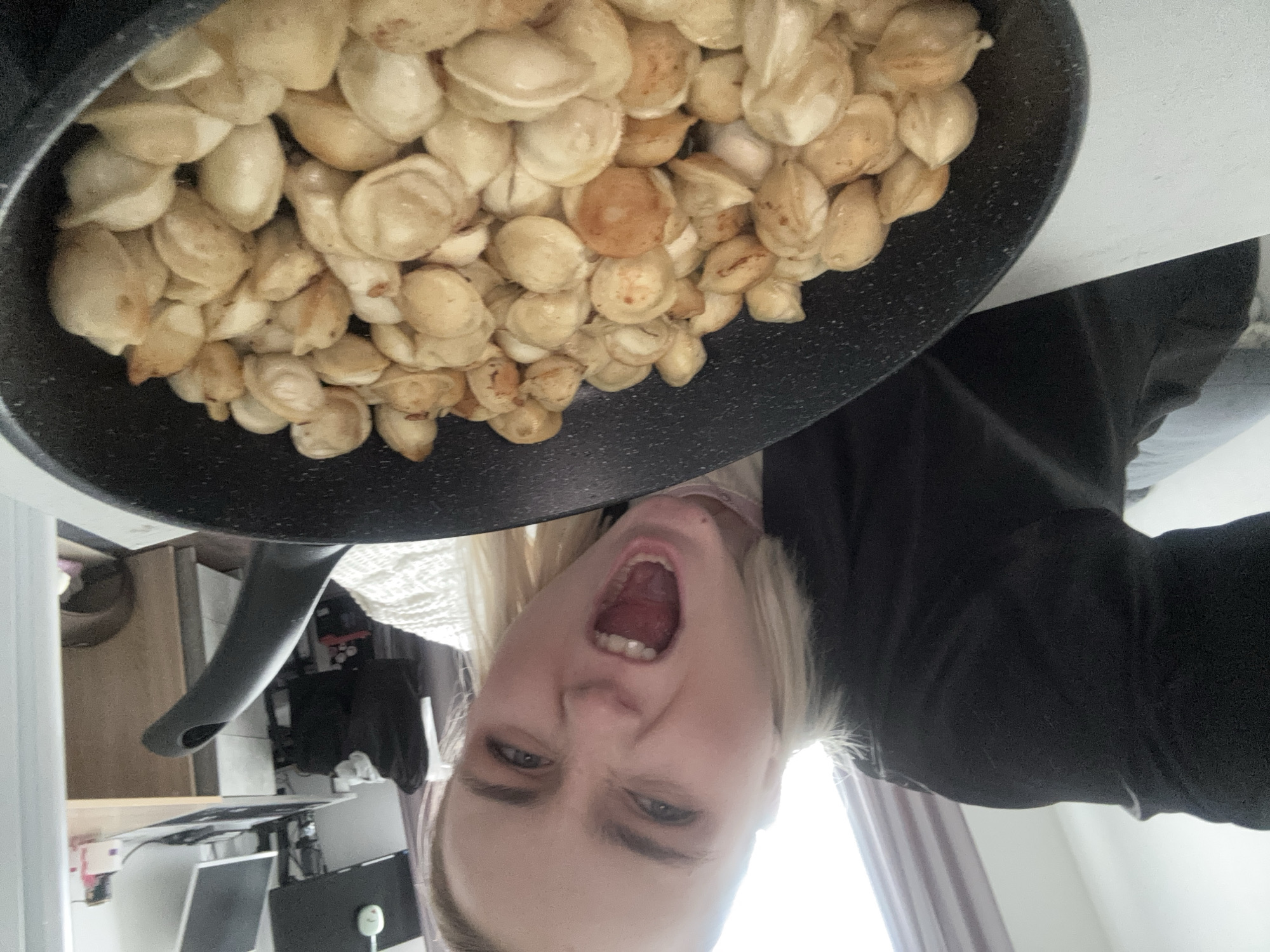 A woman is holding up a frying pan filled with dumplings. The woman has fair skin, blonde hair, and her mouth is open. She is wearing a black shirt. The frying pan is black and filled with small, cooked dumplings. The background includes a desk with computer equipment, a window, and a white wall. The specific location in Vsevolozhsk, Russia is not identifiable from the image.