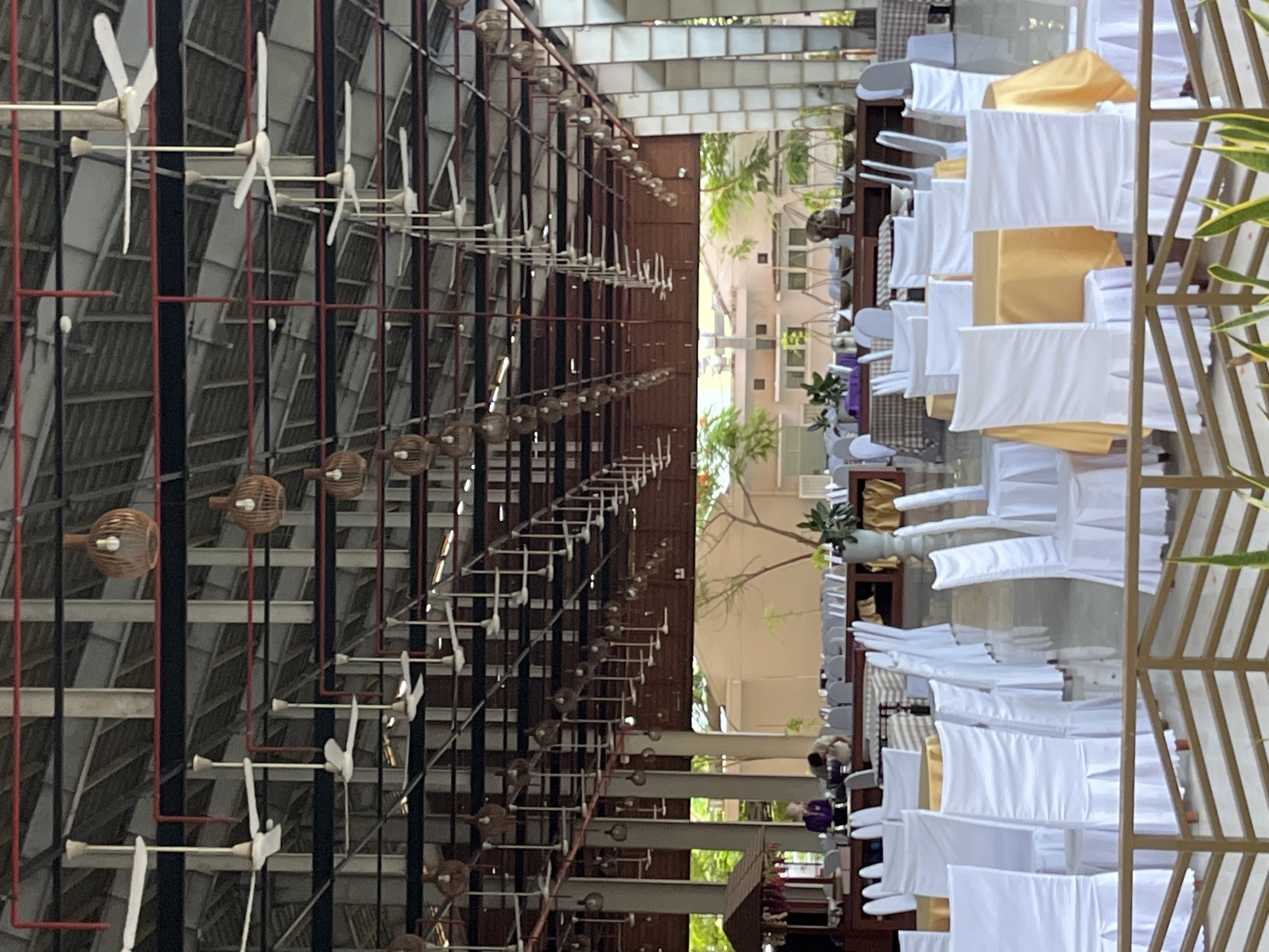The image shows an interior view of a large, open-air event space in Dong Ninh Hoa Ward, Vietnam. The space is set up for an event with numerous tables and chairs. The tables are draped with white cloths, and some are adorned with yellow tablecloths. The chairs are also covered in white fabric. Hanging from the ceiling are many electric fans and decorative lamps made of woven material. In the background, through an open wall, trees and a building can be seen. Two individuals are visible in the mid-ground, appearing to be staff or attendees. The overall impression is that of a spacious venue prepared for a gathering or celebration.