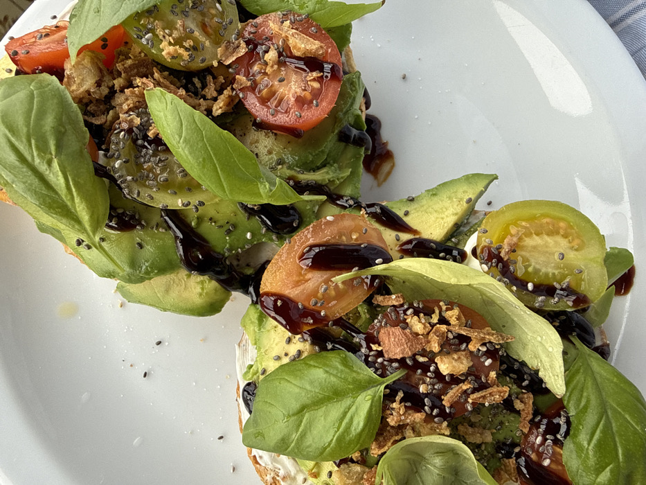Two portions of an open-faced toast or sandwich are presented on a white, glossy ceramic plate. Each piece of toast, visible beneath the toppings, is coated with a white, creamy spread. Sliced green avocado, fresh green basil leaves, and cut cherry tomatoes (featuring both red and greenish-yellow varieties) are arranged atop the spread. Black chia seeds and light brown, irregularly shaped crispy fragments are sprinkled over the entire assembly. A dark, viscous balsamic glaze is visibly drizzled across all components. A segment of blue and white striped fabric is partially visible in the bottom right corner of the frame. The image provides no discernible information to determine its specific location within Vila Nova de Gaia, Portugal.