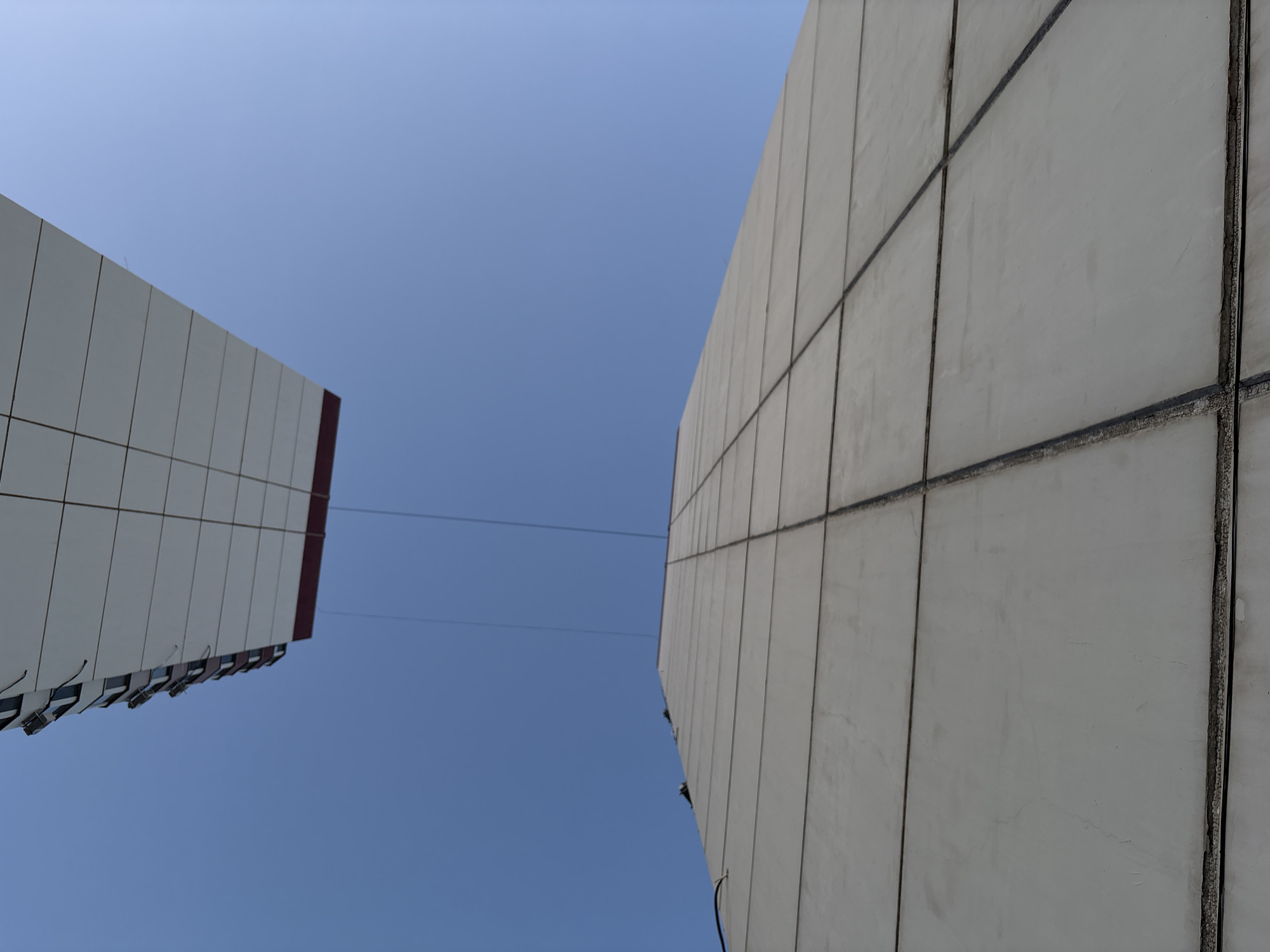 This is a low-angle shot of two concrete structures against a clear blue sky. The structure in the foreground is a large, light-colored, rectangular wall that appears to be part of a building, segmented by dark lines. The structure in the background is also rectangular and appears to be the underside of another building or overhang, with visible horizontal and vertical divisions, and a red band along its edge. Two thin wires or cables are visible stretching across the sky between the two structures, suggesting they are connected or in close proximity. The lighting indicates it is daytime, with the sun casting no visible shadows from these structures onto themselves. The composition emphasizes the geometric shapes of the architecture and the vastness of the sky. The scene is devoid of any people or other living beings.