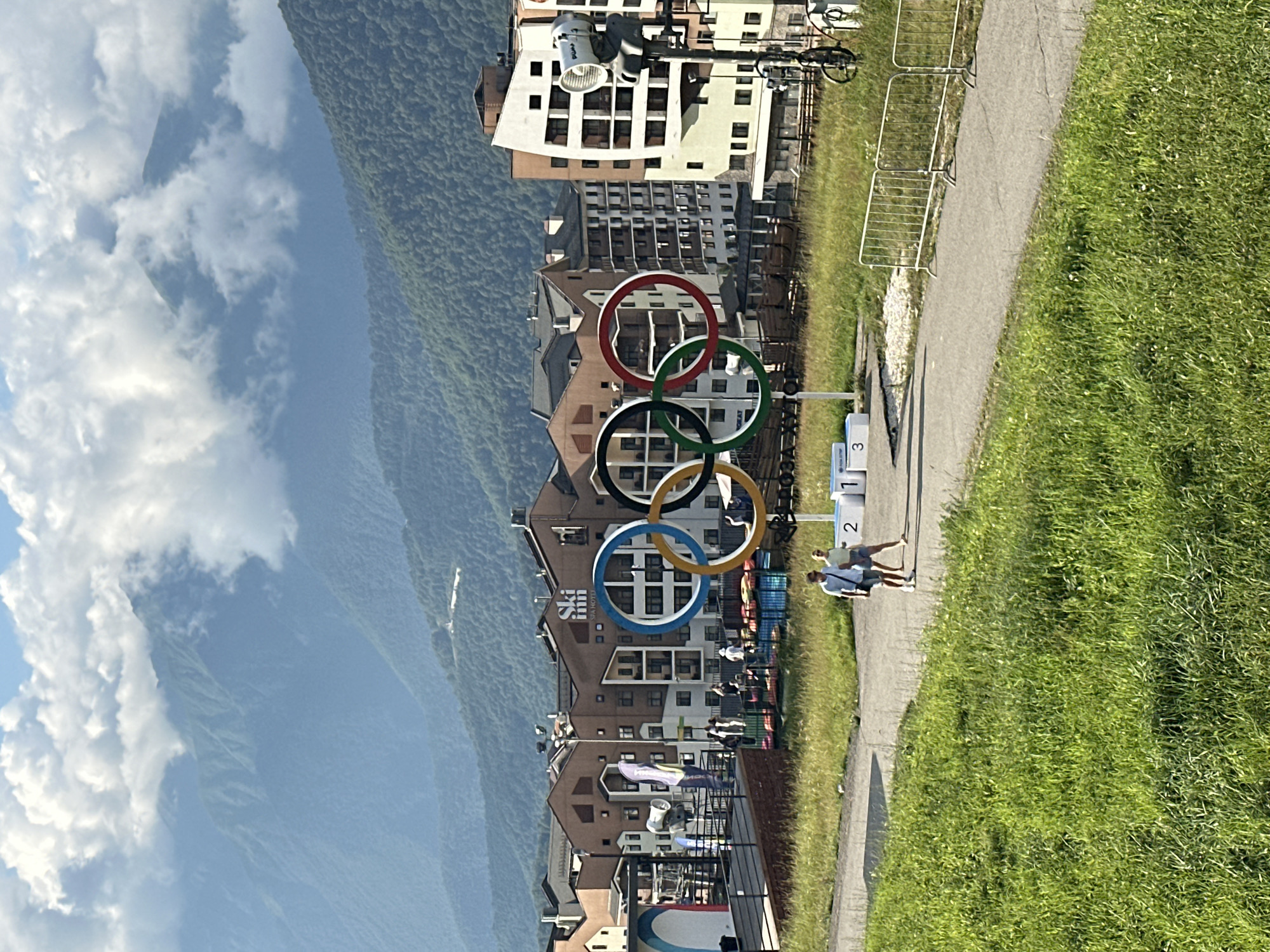 The image depicts the Olympic rings monument in Esto-Sadok, Russia, against a backdrop of lush green mountains shrouded in clouds. In the foreground, a paved path winds through a grassy area, where two people are walking. Beyond the path, the Olympic rings are prominently displayed, with the "Ski Inn Spa Hotel" building visible behind them. Several other buildings, likely hotels or apartments, are clustered in the middle ground, showcasing a blend of modern and traditional architecture. The scene is set on a sunny day, with scattered clouds adding depth to the mountainous landscape. A metal fence runs along the right side of the image, bordering the grassy area and path.