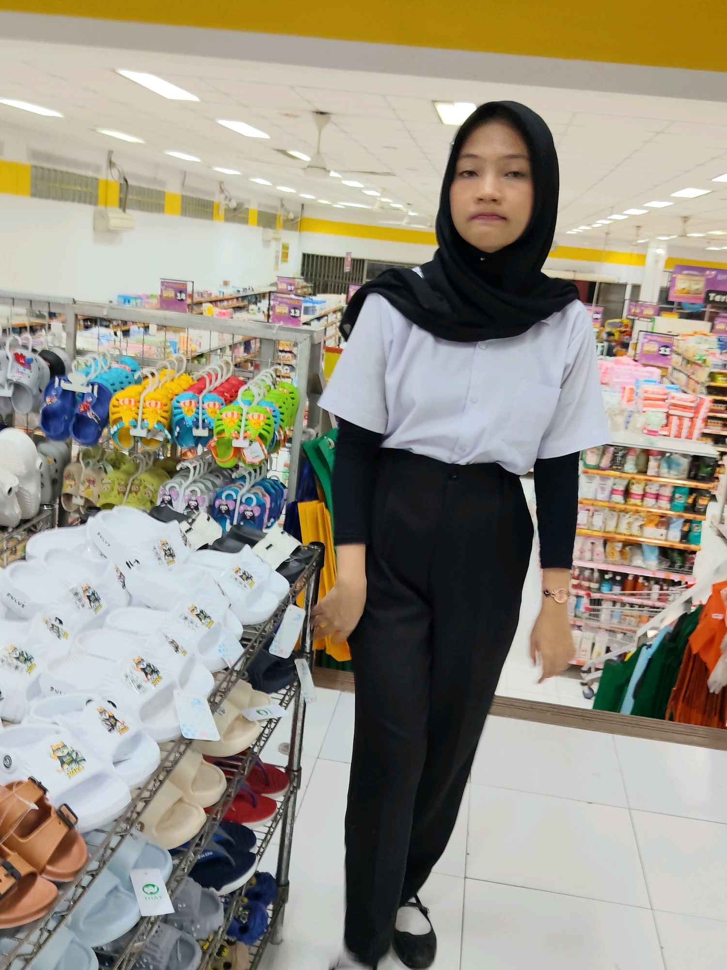 A young Indonesian woman wearing a black hijab, a light blue button-down shirt, and black pants stands in a supermarket aisle in Purwakarta, Indonesia. The aisle is filled with shelves displaying various footwear, including sandals and flip-flops. The woman is positioned in the center of the frame, looking towards the camera with a neutral expression. Behind her, shelves are stocked with products, and ceiling lights and fans are visible. The floor is tiled with white squares.
