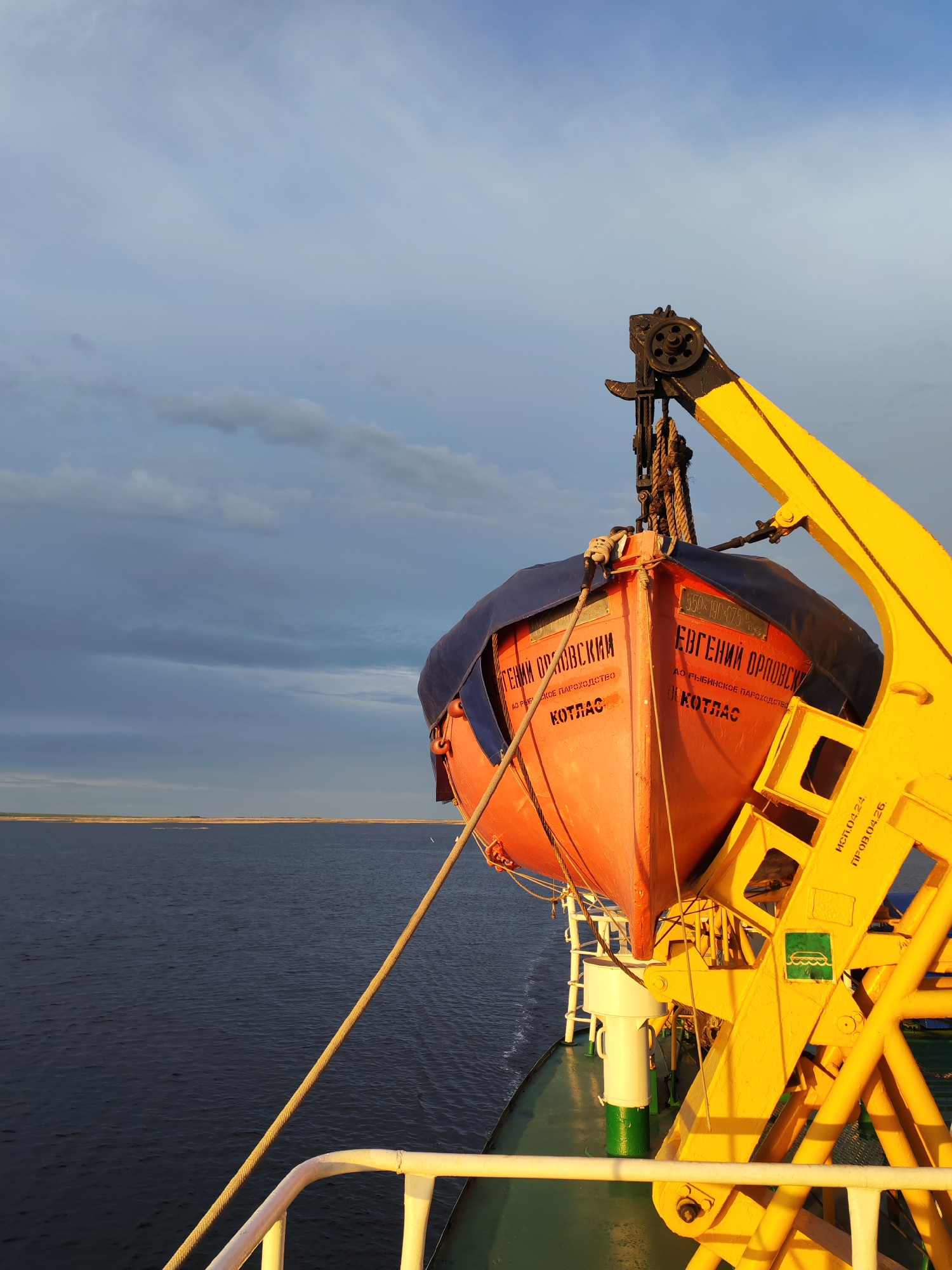 The image shows a bright orange lifeboat hanging from a yellow crane structure on a ship. The boat has blue protective covers. The text "ЕВГЕНИЙ ОРЛОВСКИЙ КОТЛАС" is printed on the side of the boat in Russian. The ship appears to be on a body of water, with land visible in the distance under a cloudy sky. The location mentioned is Bereg, Russia.