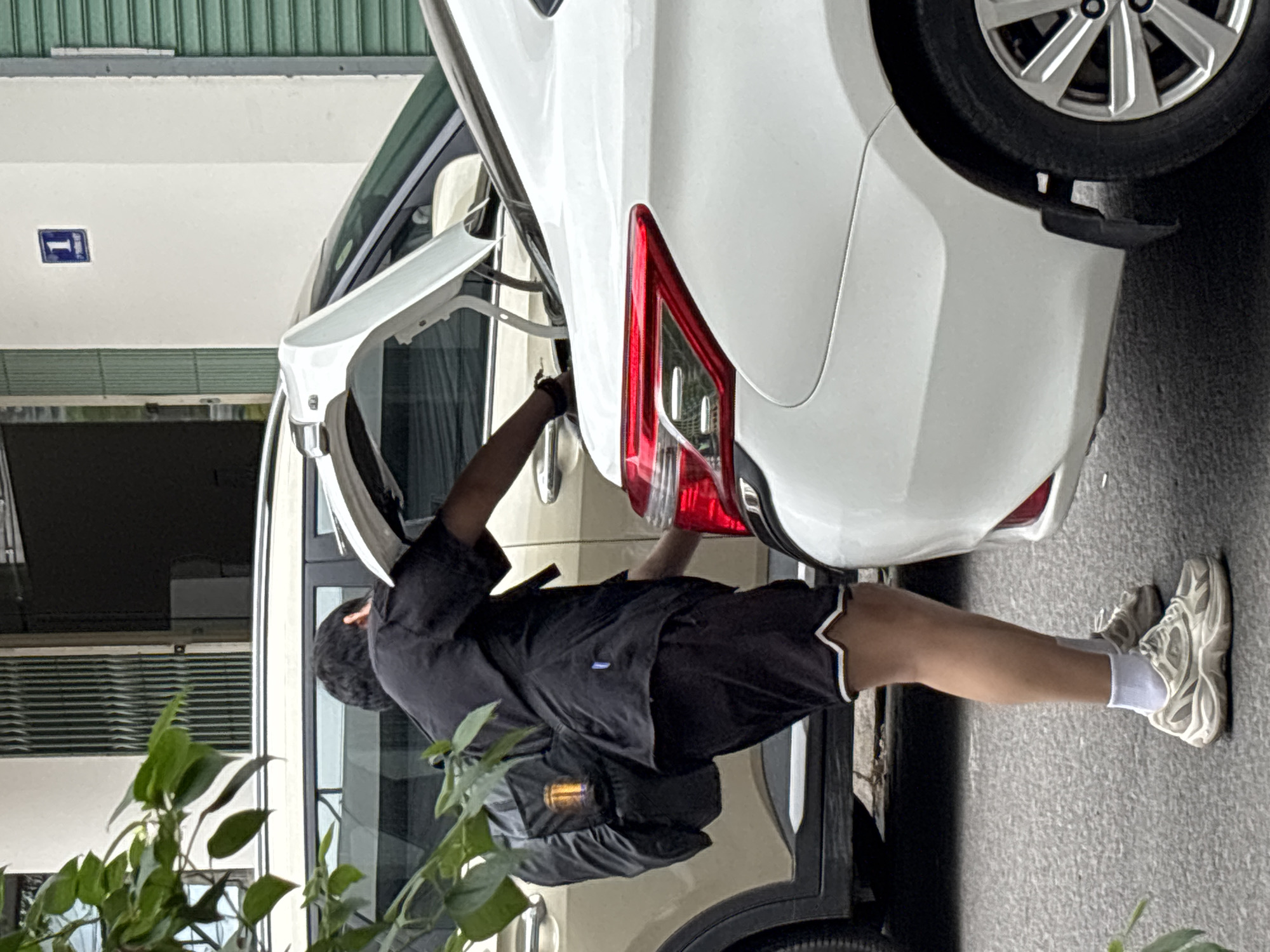 A person wearing a black t-shirt, black shorts, white socks, and white sneakers is standing by the open trunk of a white sedan. The person has a black backpack with a yellow object visible inside. They are reaching into the trunk of the white car, which is parked next to a tan SUV. The scene appears to be on a street in Nha Trang, Vietnam, indicated by the building facade with a blue sign displaying the number "1" and green metal shutters visible in the background. Green foliage is present on the left side of the image.