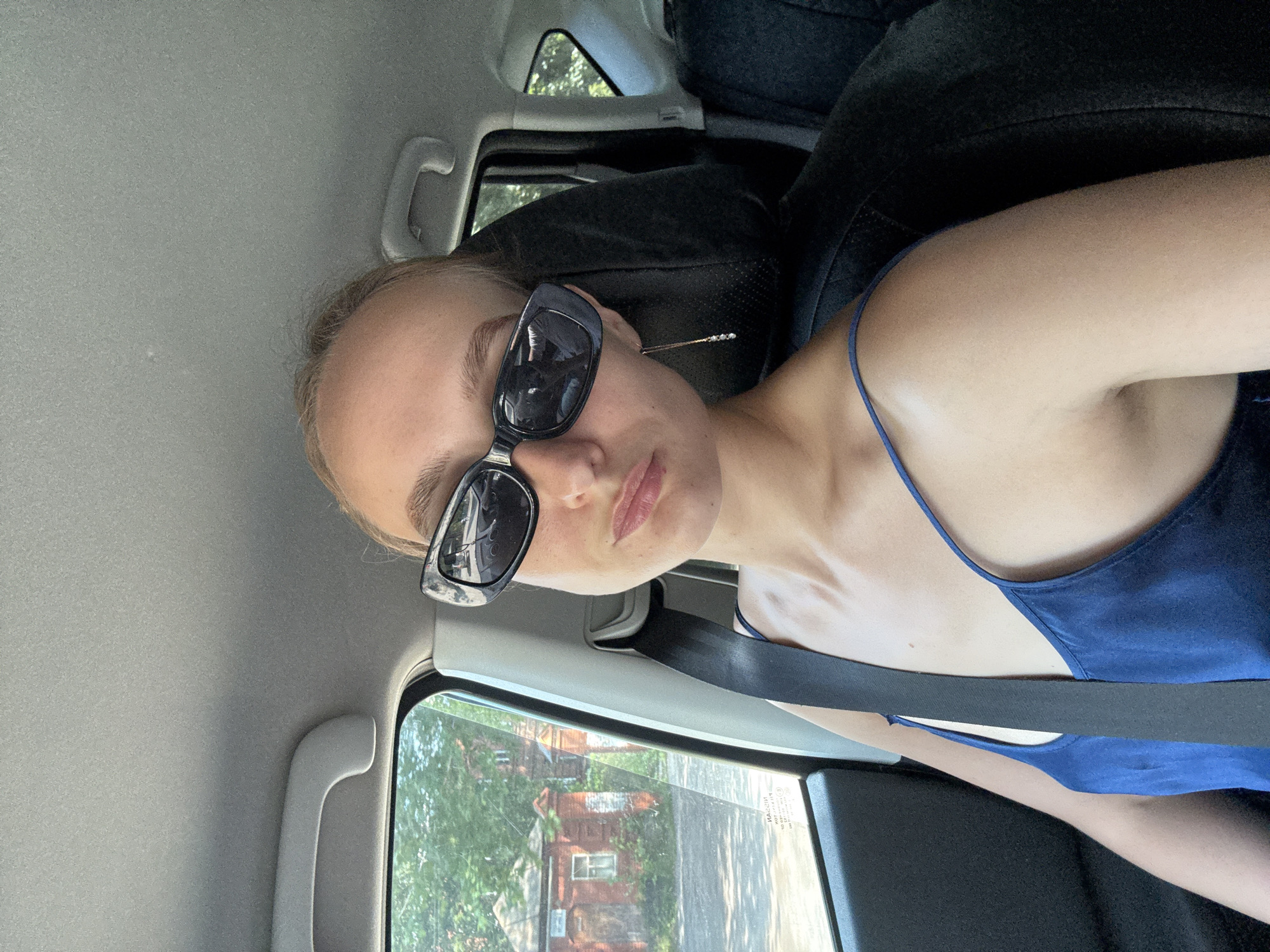 A young woman with blonde hair is sitting in the passenger seat of a car, wearing sunglasses and a blue camisole. A seatbelt is fastened across her chest. Through the car window, a brick building with a small window can be seen, surrounded by trees and foliage. The overall scene appears to be a snapshot taken during daylight hours in Votkinsk, Russia.