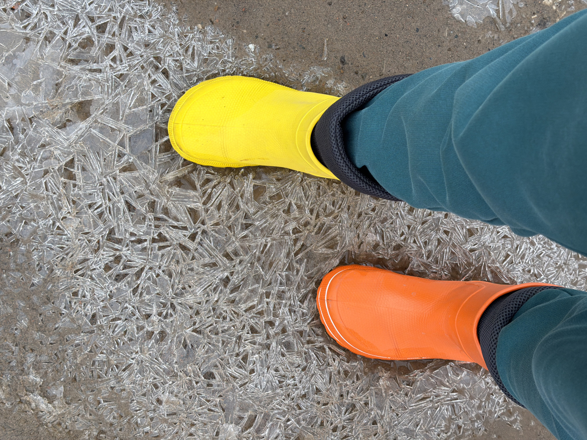 A person is standing on a layer of ice with a yellow rain boot on the left foot and an orange rain boot on the right foot. The ice has a fractured, crystalline pattern. The person is wearing teal pants tucked into the boots. The surface around the ice appears to be damp. I cannot determine the location from this image.
