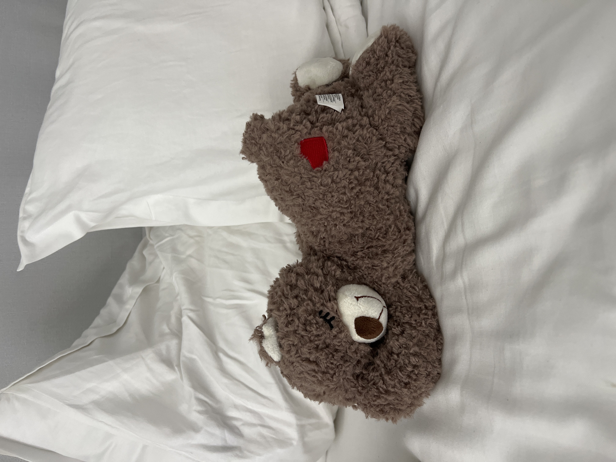 The image shows a brown, plush, stuffed bear lying on white bed linens. The bear has a red patch on its body and a white ear, and its face is visible with closed eyes. A pillow is visible behind the bear. There are no people in the scene. The location is not specified in the image and there is no reason to assume that this image was taken in Tel-Aviv, Israel.