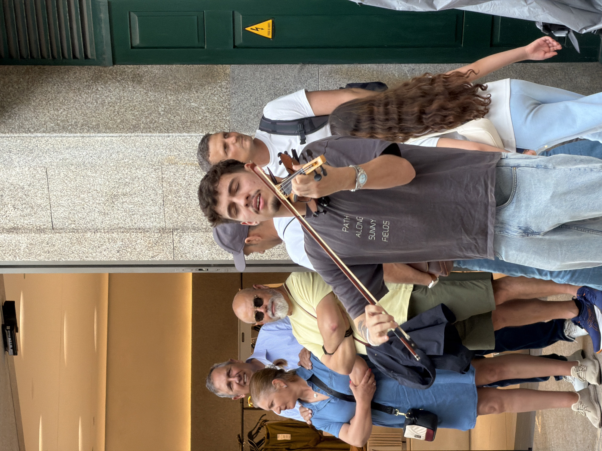 A group of people are standing near a shop in what may be the city of Porto, Portugal. A young man in a gray shirt is playing a violin. He is surrounded by other people who appear to be listening. One woman has light blue pants and a white shirt. A man behind the violinist has a white shirt and backpack. The group stands in front of a building with a green door and a yellow sign with a lightning bolt. The shop windows behind the other half of the group display merchandise.