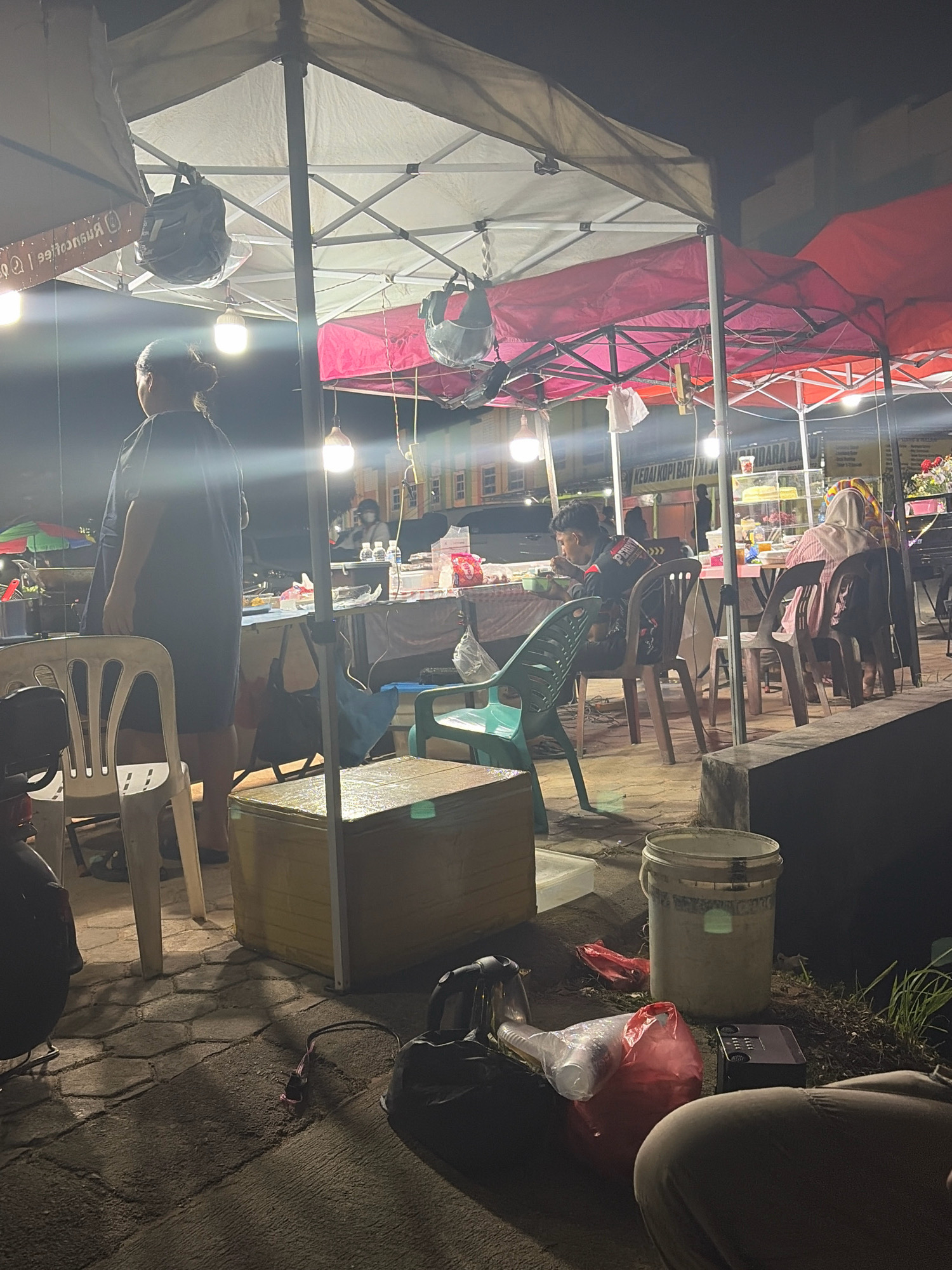 The image depicts an outdoor nighttime scene at a food stall in Tanjung Pinang, Indonesia, likely a street food area. The foreground shows a paved ground with hexagonal patterns, upon which several items are scattered, including a bucket, plastic bags with food containers, a black bag, and a black electronic device. A person's legs and torso are visible in the lower right corner, suggesting they are seated or crouched.
In the mid-ground, several stalls are set up under large tents, illuminated by bright lights. One stall features tables and chairs where people are seated. A young man wearing a black jacket with red accents is eating from a bowl. To his right, a woman wearing a hijab and a striped garment is also seated at a table. Another stall, further to the left, has a woman in a dark dress standing. Various food items and containers are visible on the tables. Several bags are hanging from the tent structures. The overall atmosphere suggests a busy, casual dining environment. The background reveals buildings and vehicles, indicating an urban setting. The signs on the stalls are partially visible, with text such as "KEDAI KOPI" suggesting a coffee shop or food stall.