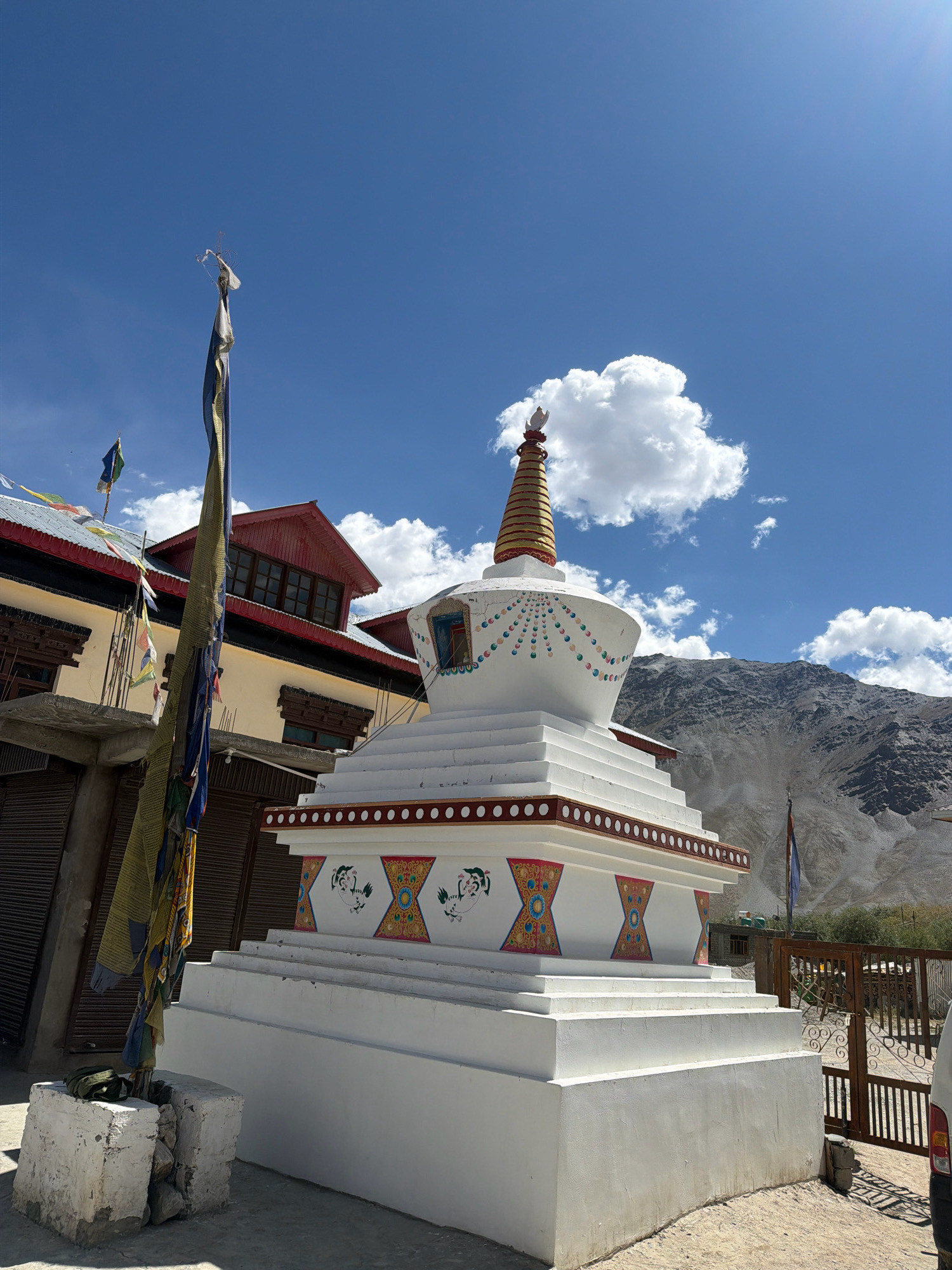 The image shows a Buddhist stupa in Padum, India, with a monastery or guesthouse in the background. The stupa is white with a tiered base and a conical top adorned with gold and red bands. Prayer flags, in various colors and tattered from the wind, hang from a pole to the left of the stupa. The monastery features a yellow facade with red trim and dark wooden window frames. Behind the stupa, a barren, rocky mountain range rises against a bright blue sky with scattered white clouds. A wooden gate and a fence are visible to the right of the stupa. The scene is bathed in sunlight, casting shadows on the ground. There are no people visible in the frame.