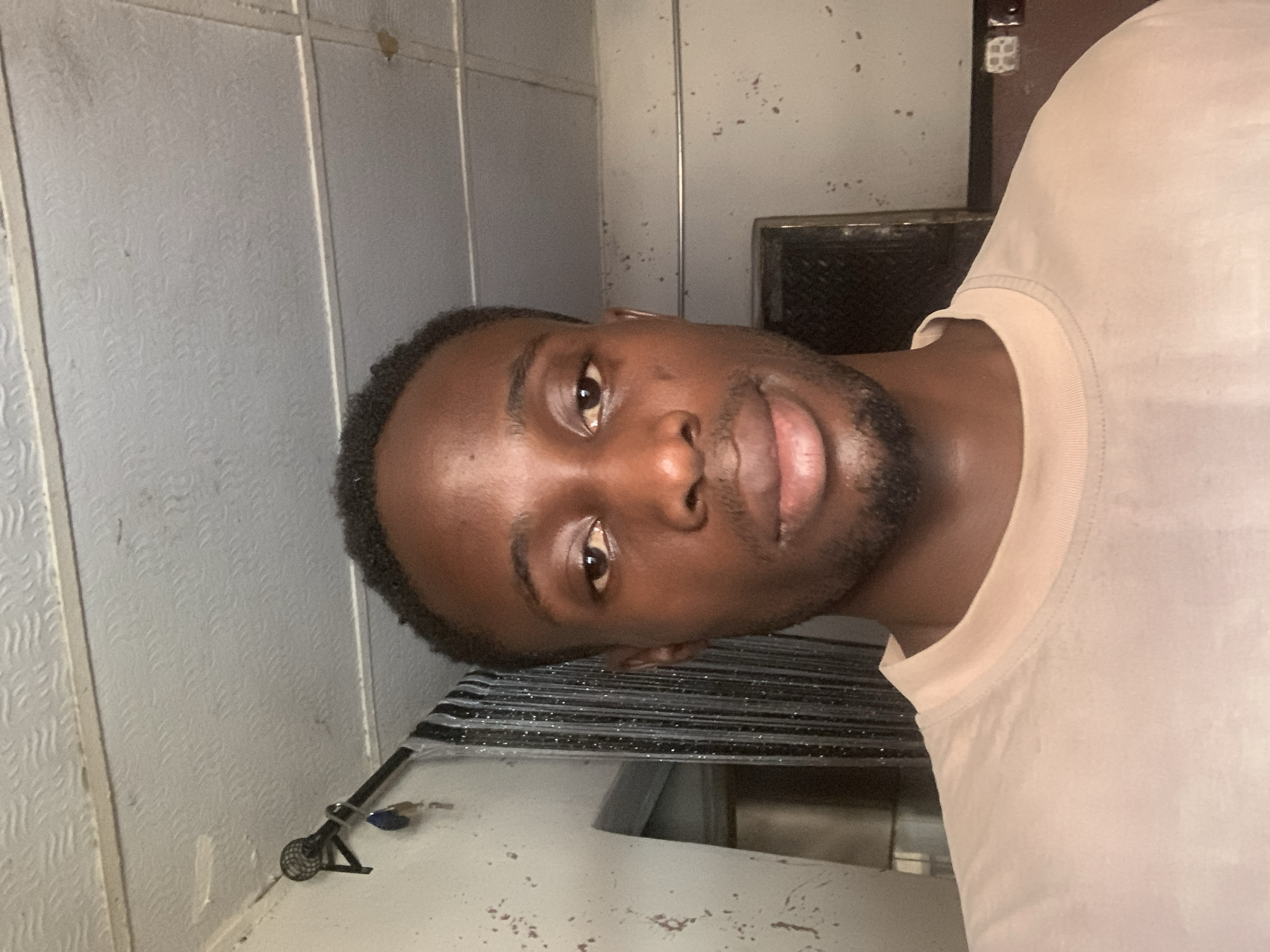 A portrait-style image, rotated ninety degrees clockwise, features a young Black man looking directly into the camera. He has dark, short curly hair and a neatly trimmed beard. His expression is neutral with a slight hint of a smile. He is wearing a plain, light-colored (likely beige or light pink) crew-neck t-shirt.
The background indicates an indoor setting, possibly a room or hallway. The wall behind him is light gray or off-white, characterized by a subtle textured pattern and vertical seams, suggesting panels or tiles. Visible wear and tear, including chipped paint and discoloration, are present on the wall surfaces. To the viewer's lower left (when the image is viewed upright), a dark, metallic-looking fixture with a small key or object hanging from it is partially visible. Below his chin, a dark, horizontal slatted structure, possibly a window blind or a grate, is present. Further to the upper right, a white electrical outlet or switch is visible against a darker background. The lighting appears to be artificial and somewhat dim. No text is discernible in the image. The overall impression is a casual, personal photograph. Given the context, this scene is from Lokoja, Nigeria.