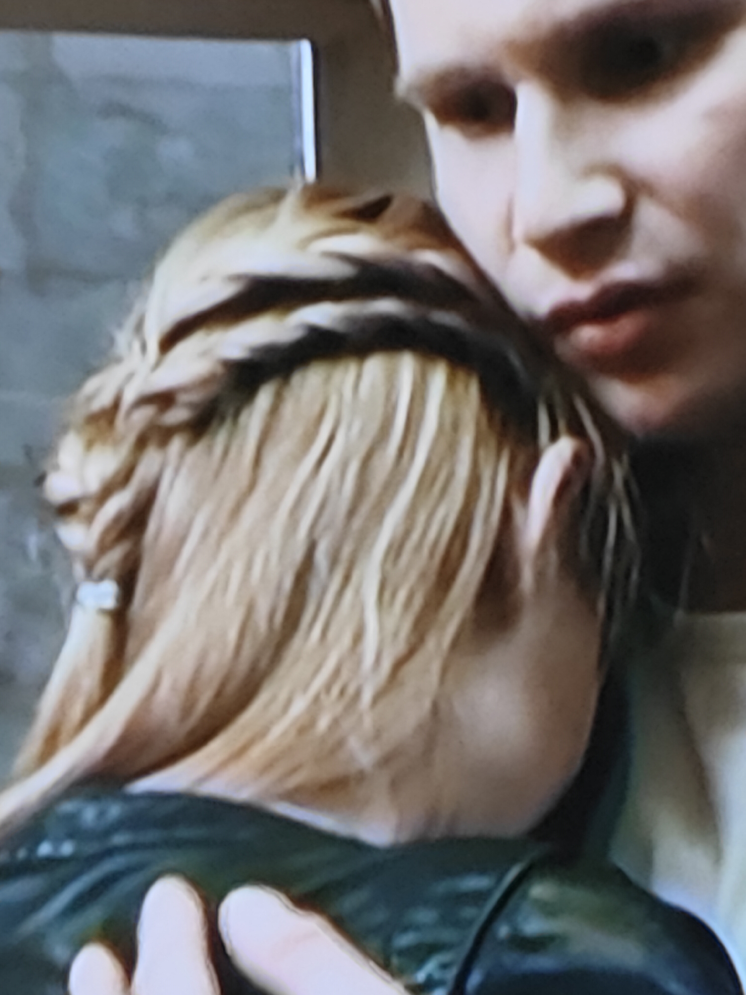 The media file is a close-up, grainy, and slightly pixelated image of two people. The foreground subject is a person viewed from the back and side, with blonde hair styled in braids and loose strands. They are wearing a dark, possibly leather, jacket. The second subject, a person with light-colored hair and fair skin, is visible in the background, leaning in and embracing the foreground subject. Their face is partially visible, showing their nose, mouth, and chin, with a neutral or slightly pensive expression. The lighting suggests an indoor setting, possibly dimly lit, with no obvious time of day or weather cues. The background is indistinct, with what appears to be a wall with a textured surface and a framed opening or window. There is no visible text in the image. The overall mood is intimate and perhaps somber, given the close embrace and the lighting.