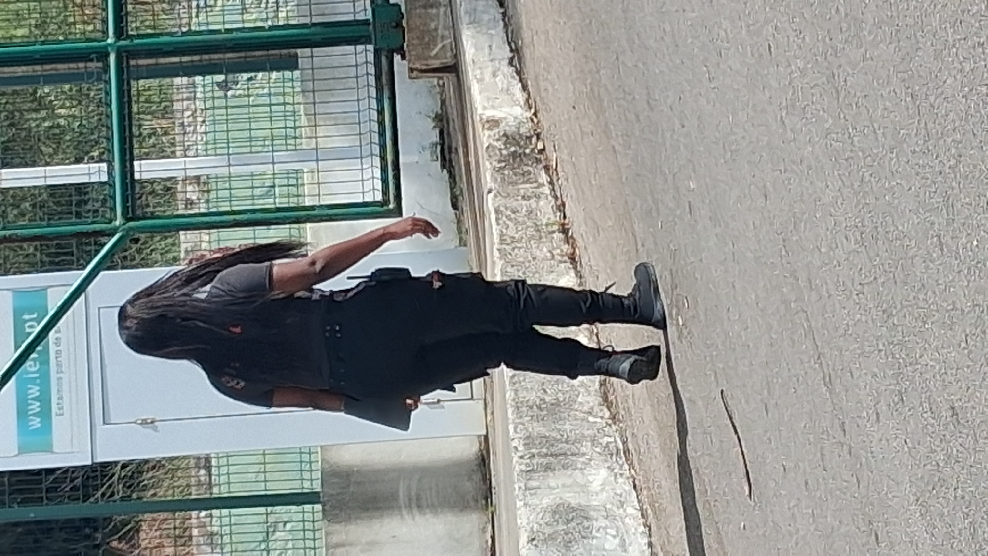 A person with long dark hair, wearing a black t-shirt and black pants with tactical gear, is standing on a paved surface. The person is seen from the side, with their left arm extended slightly. They appear to be holding a dark object in their left hand. The scene is outdoors, with a green metal fence in the background. Behind the fence, there is a building with a white door and a sign that has text on it. The ground is a rough, gray asphalt. The lighting suggests it is daytime, with shadows visible on the ground. The image is oriented vertically, with the top of the frame showing the top of the fence and the bottom showing the pavement. The provided crops offer closer views of the person's clothing, hair, and feet, as well as the sign on the building and the background fence. The text on the sign is partially visible and appears to be "www.ie.pt" and below it "Estamos perto de". The overall impression is a candid shot of someone in uniform or tactical gear standing near a building in an outdoor urban or semi-urban environment.