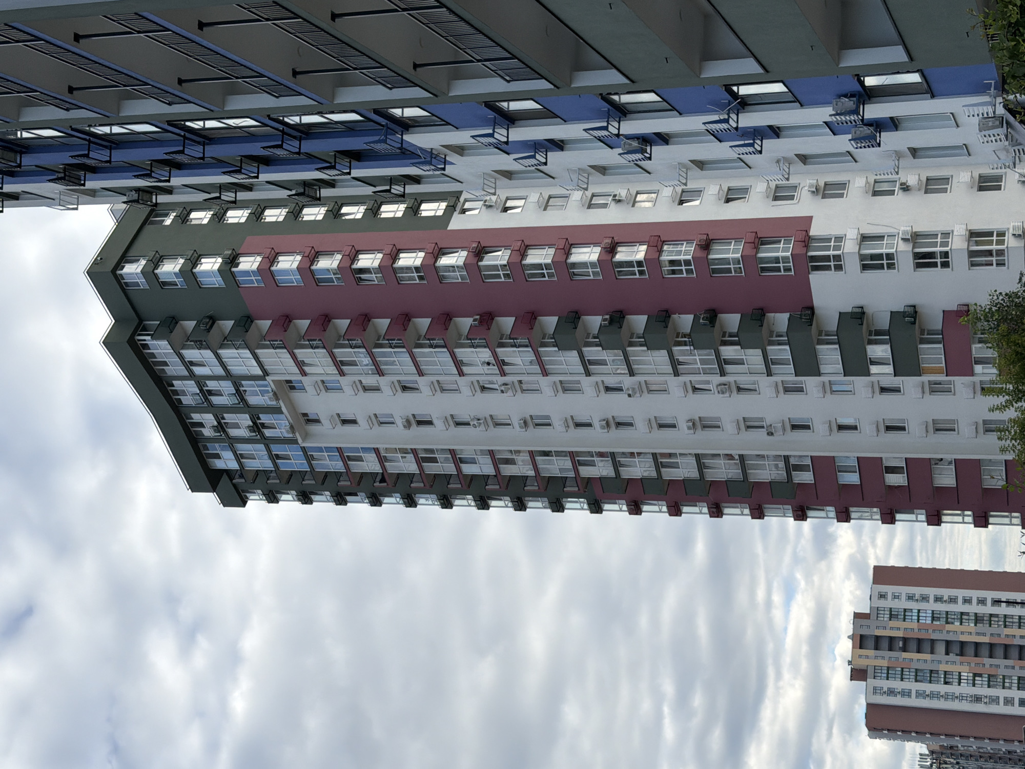 The image depicts a section of a modern residential complex in Kyiv, Ukraine, under a cloudy sky. The dominant feature is a tall, multi-story apartment building with a segmented facade. The building's exterior is characterized by a combination of white, dark green, and a distinctive maroon color, accentuating different sections and balconies. Numerous windows, many with protruding balconies, punctuate the facade. Several units have air conditioning units mounted externally.
To the right of this primary building, another apartment building is visible, partially obscuring it. This adjacent structure features a blue and white color scheme, also with numerous windows and balconies. Its balconies are marked by dark railings.
In the background, to the left, a smaller, lower-rise apartment building is visible. This building has a more colorful facade, with horizontal bands of beige, grey, and orange.
The overall impression is of a newly constructed or recently renovated urban residential area, characterized by vertical living structures. The lighting suggests it is daytime, with diffused light due to the overcast conditions. No people are discernible in the image.