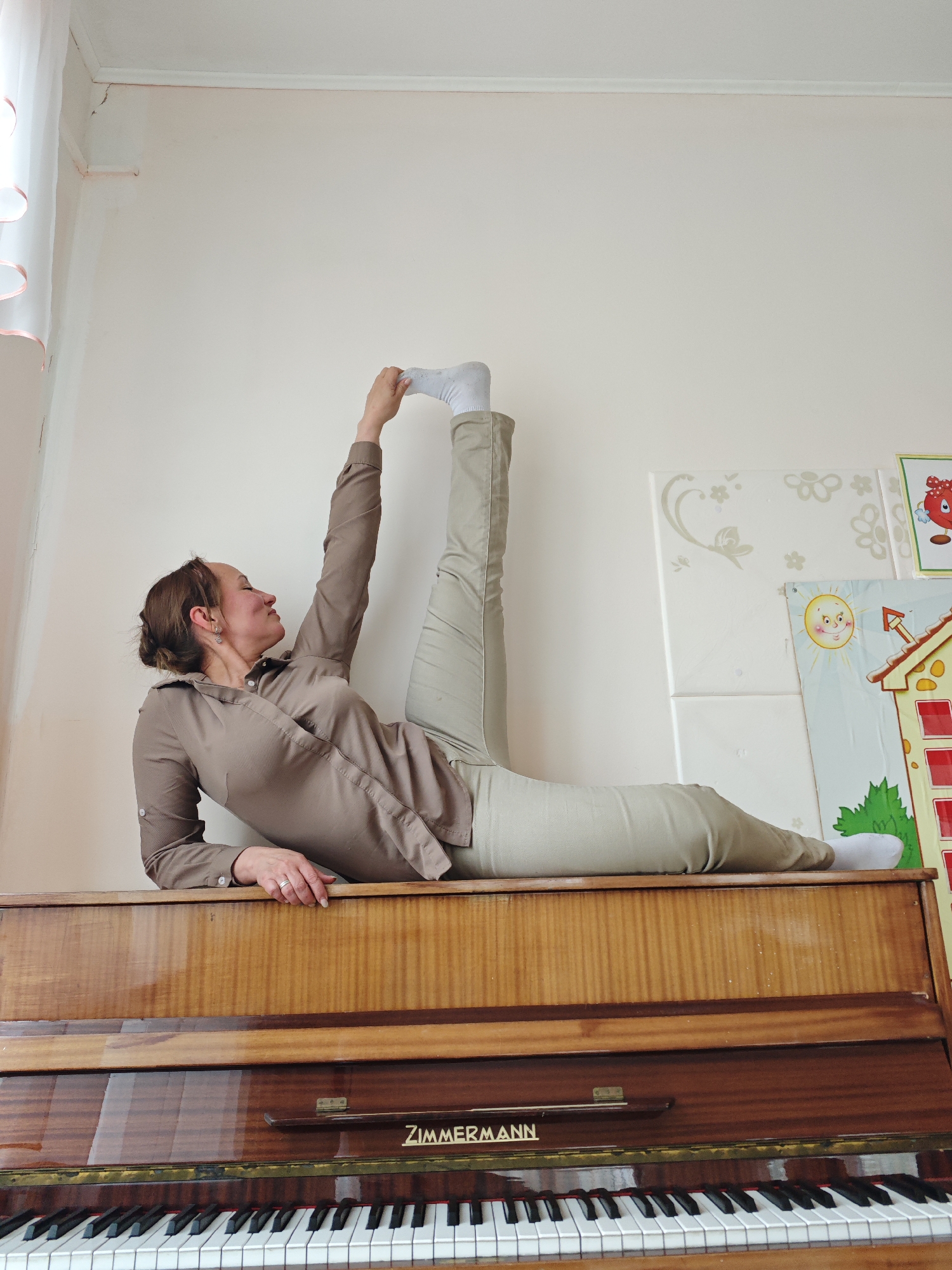 A woman is lying on her back on top of a piano, extending one leg straight up in the air and grasping her foot with her hand. She is wearing a long-sleeved brown shirt and khaki pants. She is smiling and appears to be enjoying the activity. The piano is a dark wood color with the name "Zimmermann" displayed on it. The background consists of a white wall with some decorations. The location is Markovskii, Russia.