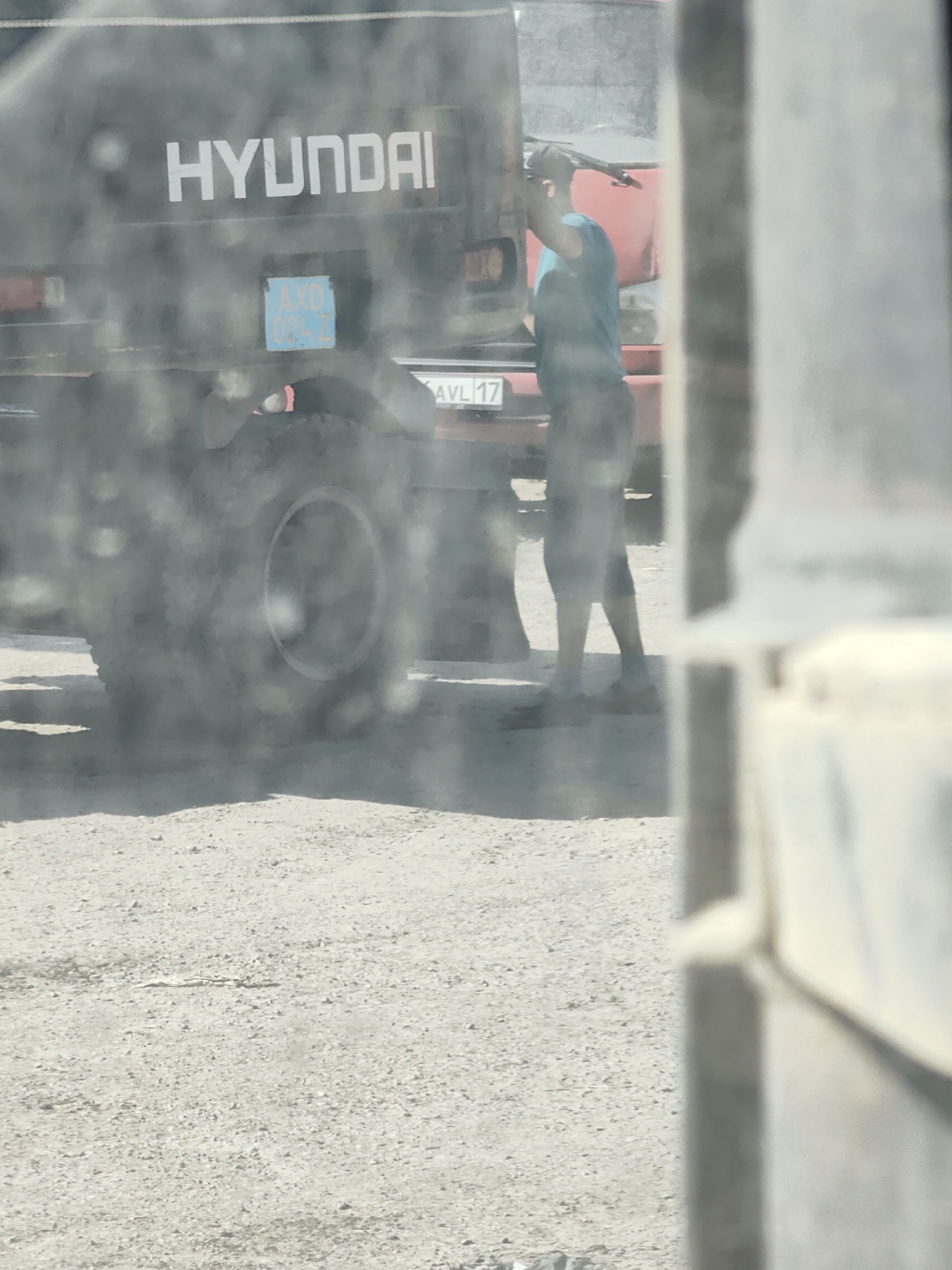 A person is standing near the back of a dark-colored truck, possibly a Hyundai, with their arm raised as if working on it. A license plate with the characters "AXD 034 Z" is visible on the truck. Another license plate, "AVL 17," is also visible on the truck. The truck is on a gravelly surface. The image appears to be taken through a dirty window or windshield, with significant blur and dust. The background shows another red truck. The scene is outdoors in what appears to be a dry, sunny environment. Based on the license plate format and general appearance, this scene is likely in Shymkent, Kazakhstan.