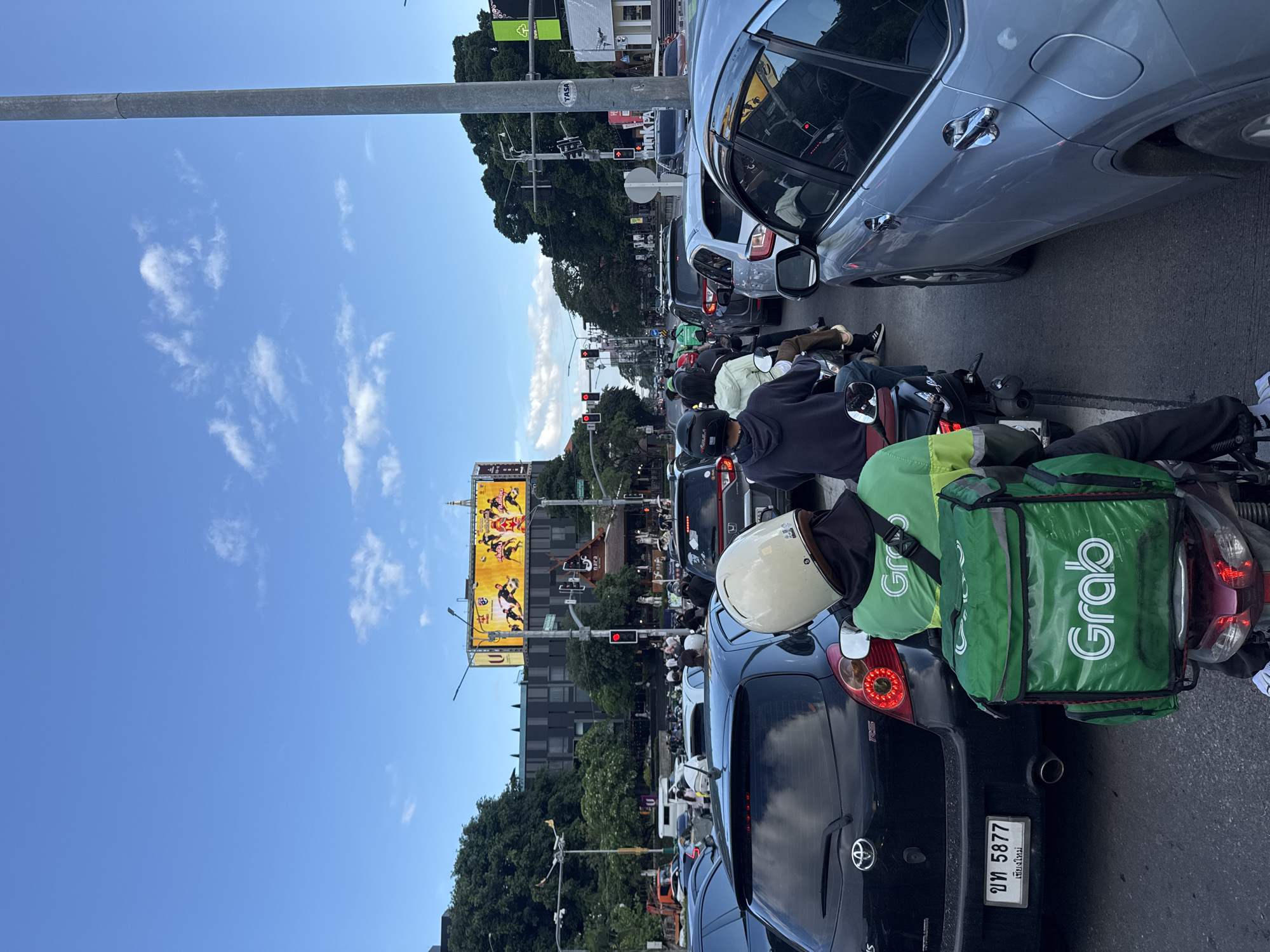This is a street-level photograph taken from behind a motorcycle rider in a traffic jam on a clear day. The rider is wearing a white helmet and a green Grab delivery uniform with a large green Grab bag on the back of their motorcycle. The motorcycle is positioned in the middle lane of a multi-lane road. In front of the motorcycle, a black Toyota hatchback is visible, showing its rear bumper, taillights, and a license plate that reads "บท 5877" and "เชียงใหม่" in Thai script, indicating the location is Chiang Mai, Thailand. To the right of the black car, a silver SUV is in the adjacent lane. The road is congested with numerous other cars and motorcycles. Overhead, a large digital billboard displays a yellow advertisement featuring several individuals in athletic attire. Traffic lights are visible, some showing red. Trees line the sides of the road, and buildings are present in the background, including one with a dark facade. The sky is blue with scattered white clouds. The scene depicts a typical urban traffic situation in Chiang Mai.
