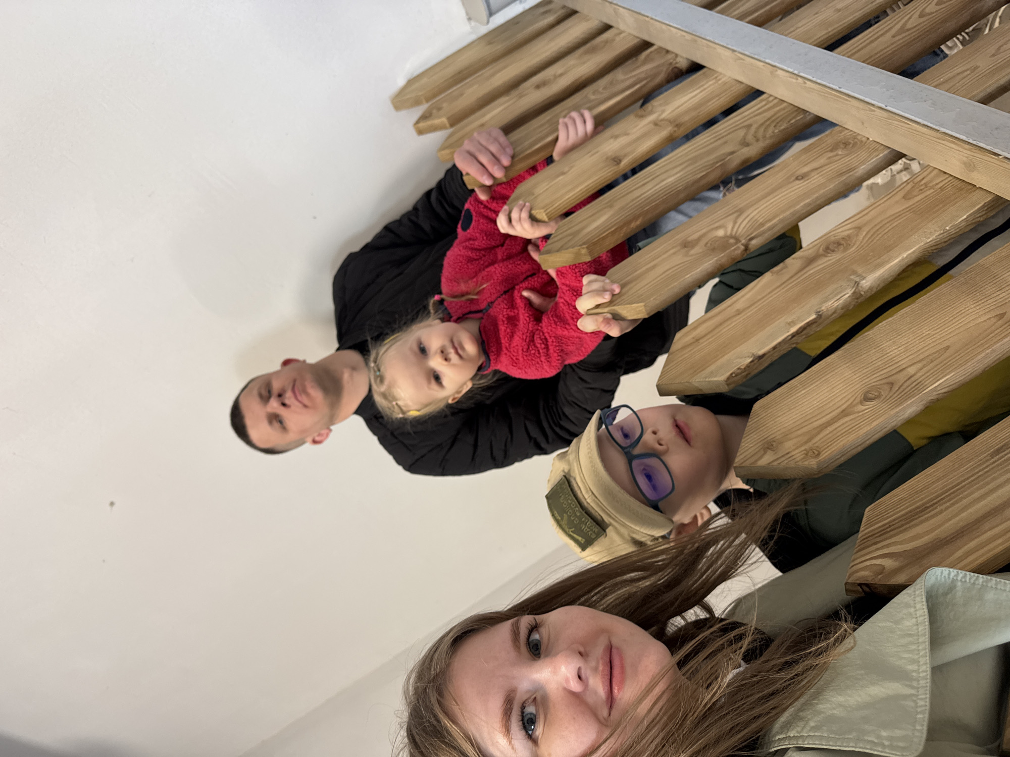 A selfie, oriented vertically, shows a family of four underneath a wooden structure. In the foreground, a woman with long brown hair looks into the camera. Behind her, a boy wearing glasses and a cap looks up. Behind them, a man wearing a black jacket holds a young girl in a red fleece jacket beneath the wooden structure. The structure consists of evenly spaced wooden beams. The location is Perm, Russia.