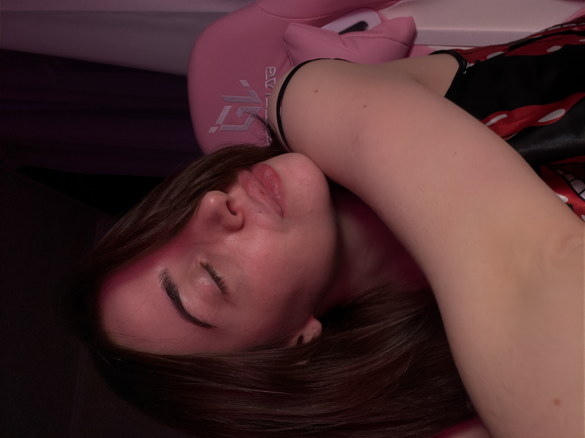 A young woman with brown hair and closed eyes is leaning her head on her arm. She is wearing a black top with a red and white pattern. She is sitting in a pink chair with a logo on the back. The lighting in the image is dim and appears to be from a pink light source, casting a reddish hue on her face and arm. The background is dark, with some hints of purple curtains.