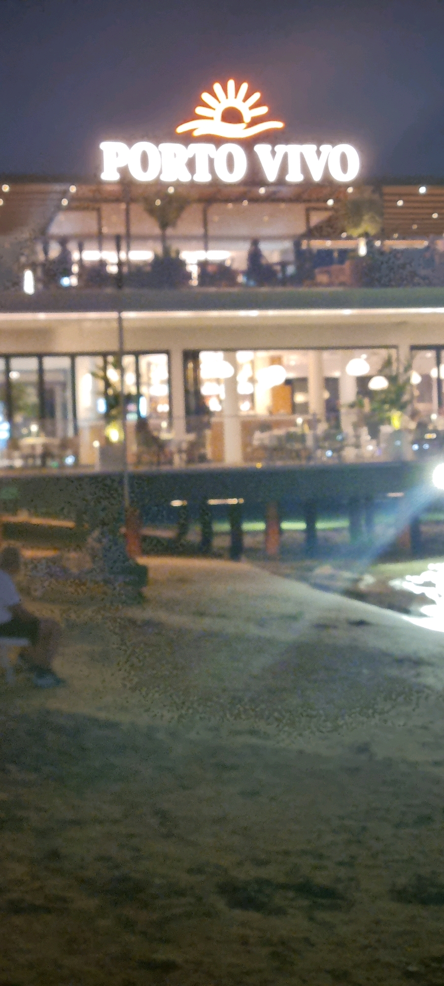 This is a nighttime outdoor shot of a multi-level establishment identified by a lit sign reading "PORTO VIVO" with a stylized sun logo above it. The building appears to be a restaurant or bar with outdoor seating on multiple levels, indicated by tables and patrons visible through glass walls and on open-air balconies. The overall lighting suggests an evening or nighttime setting, with artificial lights illuminating the building and its surroundings.
In the foreground, a paved area leads to a grassy or sandy expanse. A person is seated on a bench on the left side of the frame, facing away from the camera, seemingly observing the scene. The background includes a dark sky. The building is situated in a location that appears to be near a waterfront or a body of water, given the reflection of lights on the ground in the lower right quadrant of the image. The architecture suggests a modern design.
The establishment is located in Bashkia Durrës, Albania.