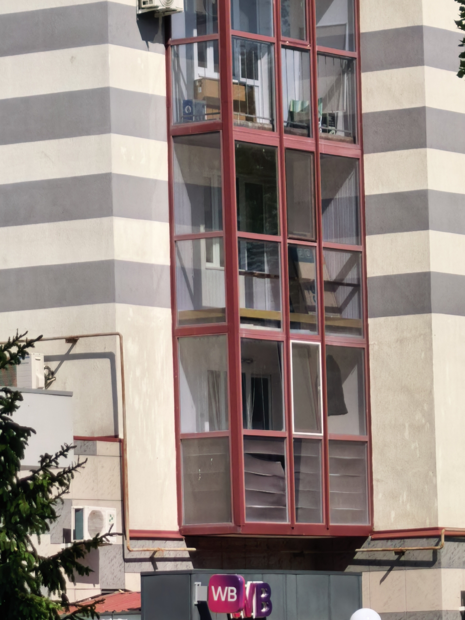 The image shows a tall, narrow exterior of a building, possibly located in the city of Sterlitamak, Russia, based on the Cyrillic lettering "WB" on a sign at the bottom of the frame. The building features a distinctive vertical window section with a red frame, comprised of multiple glass panels across its height. The primary facade of the building is patterned with horizontal gray stripes. An air conditioning unit is mounted on the wall near the top of the framed image. Reflections within the window panes indicate interior elements, including furniture and window blinds. Additionally, a tree branch is visible in the lower left corner of the frame. An additional building with an air conditioning unit is also visible, but further away than the main building. A rusty pipe is attached to the buildings facade. The sign at the bottom of the frame also includes purple and blue colors. The overall lighting suggests a sunny day.