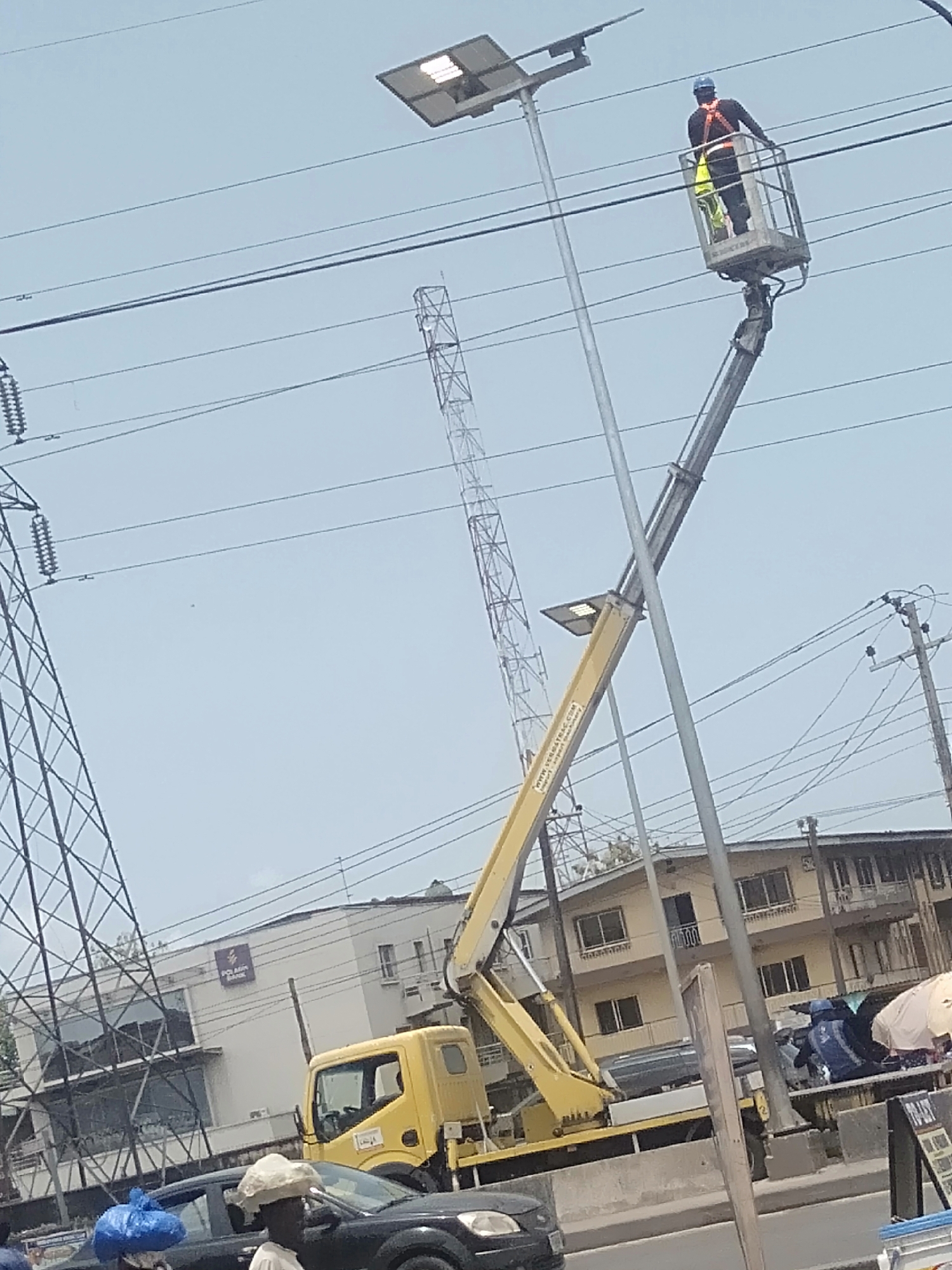 In Shangisha, Nigeria, a maintenance worker wearing a safety harness and a blue hard hat is positioned in the elevated bucket of a yellow aerial lift truck. The truck is parked on a street lined with buildings, including a building with the signage "POLARIS BANK." The worker appears to be servicing or inspecting a solar-powered street light. The aerial lift is extended upward, placing the bucket at the height of the street light. Overhead power lines stretch across the scene, and in the background, a tall transmission tower is visible. Street-level activities include passing vehicles and pedestrians.