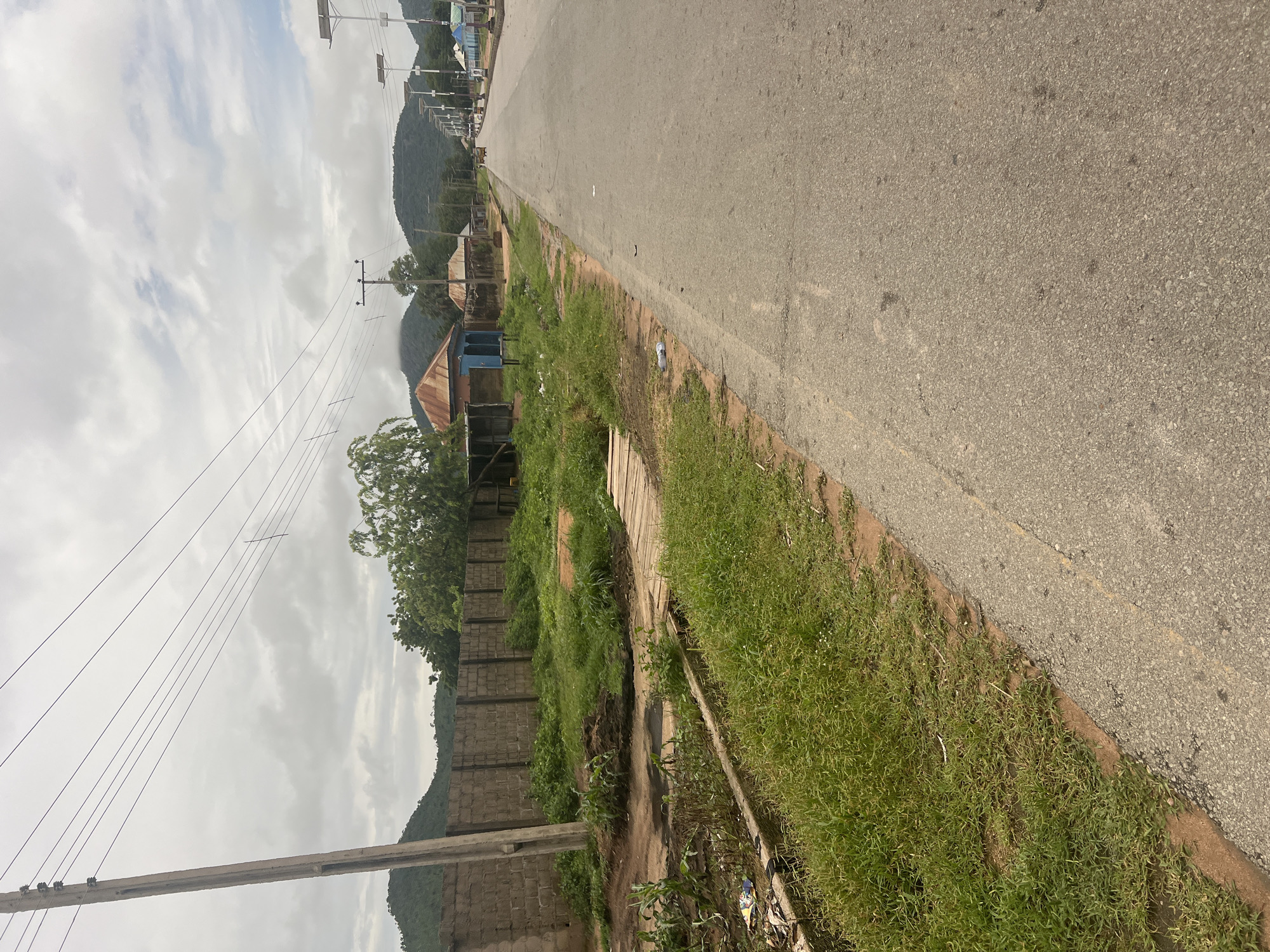 The image depicts a rural road in Kona Garu, Nigeria, under a cloudy sky. The scene is a roadside view, showing a paved road on the right side of the frame that stretches into the distance. To the left of the road, there is a strip of overgrown grass and dirt, bordered by a low concrete wall. Beyond the wall, several structures with corrugated metal roofs are visible, suggesting a small settlement. In the background, rolling hills covered in vegetation rise up, partially obscured by the overcast sky. Electrical poles and wires are strung along the side of the road, indicating the presence of electricity infrastructure. The overall impression is one of a quiet, everyday scene in a developing area, with no people or specific activities immediately apparent. The lighting suggests it is daytime, but the cloud cover makes it difficult to determine the exact time.