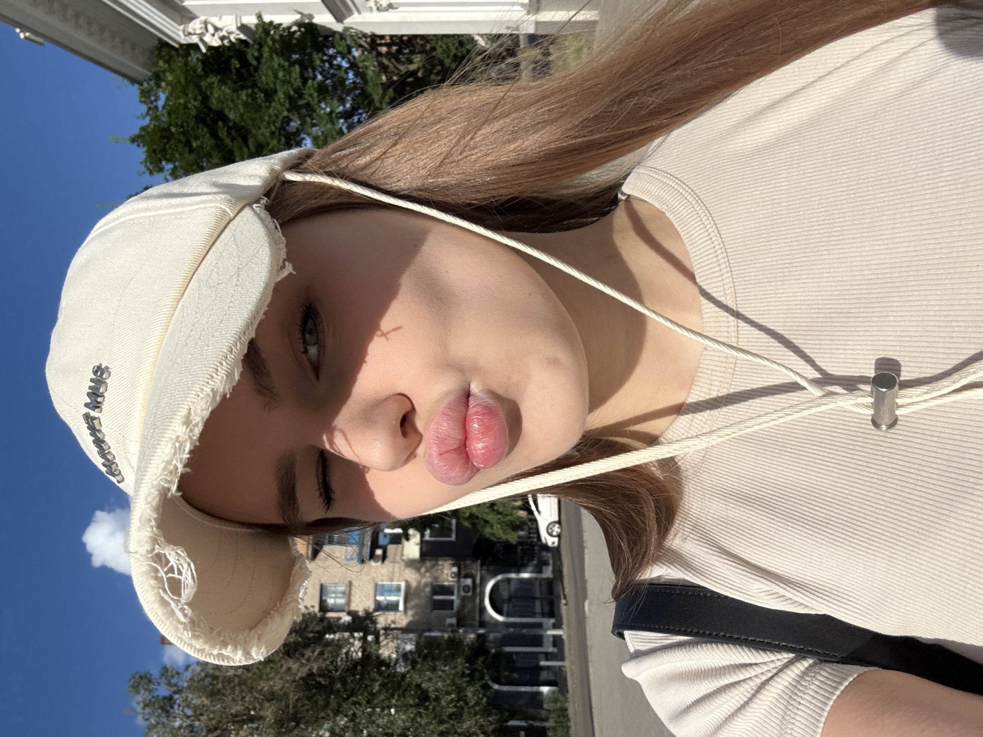 A young person, likely female given the long blonde hair, is pictured in a close-up, selfie-style shot. They are wearing a light-colored bucket hat that has frayed edges and some visible stitching. The hat has text printed on the front in a reflective, metallic font that appears to read "MARKET MUS". They are also wearing a ribbed, light beige top.
The person's face is the main focus, with their lips pursed in a kissing or pouting gesture. One eye is partially closed, while the other is open and looking towards the camera. Sunlight casts a shadow across their face. The overall expression could be interpreted as playful or coy.
The background suggests an outdoor setting on a sunny day. Visible elements include a bright blue sky with a few white clouds, lush green trees, and part of a building with architectural details, possibly a white ornate structure on the top left. Further in the background, there are residential buildings and a white car, providing context of an urban environment. The lighting indicates it is daytime, likely mid-morning or afternoon due to the strong sunlight and shadows. The orientation of the image is rotated 90 degrees counter-clockwise. Based on the location context provided, this scene is likely in Dnipro, Ukraine.