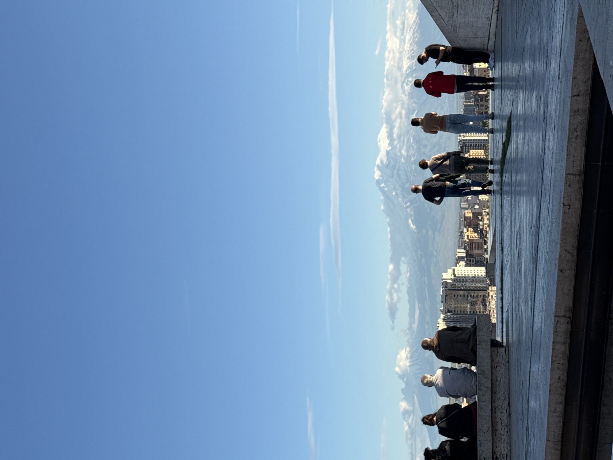 A low-angle, outdoor, full-shot photograph shows a group of people, who are standing on a stone ledge overlooking a mountain range and city buildings under a clear, blue sky.
The right side of the photograph contains a group of nine people. They are standing near the edge of a stone ledge, and are leaning over looking at the scene below. They are of various heights and are wearing various types of clothing.
Below the ledge, city buildings with beige, white, and tan-colored exteriors can be seen. These buildings gradually transition into the base of a mountain range. The mountains are capped in snow, and have some low-lying clouds near the base.
The sky is light blue in color and occupies most of the left side of the frame. The entire image is taken during the day.
The image may be of the Cascade monument in Yerevan, Armenia.