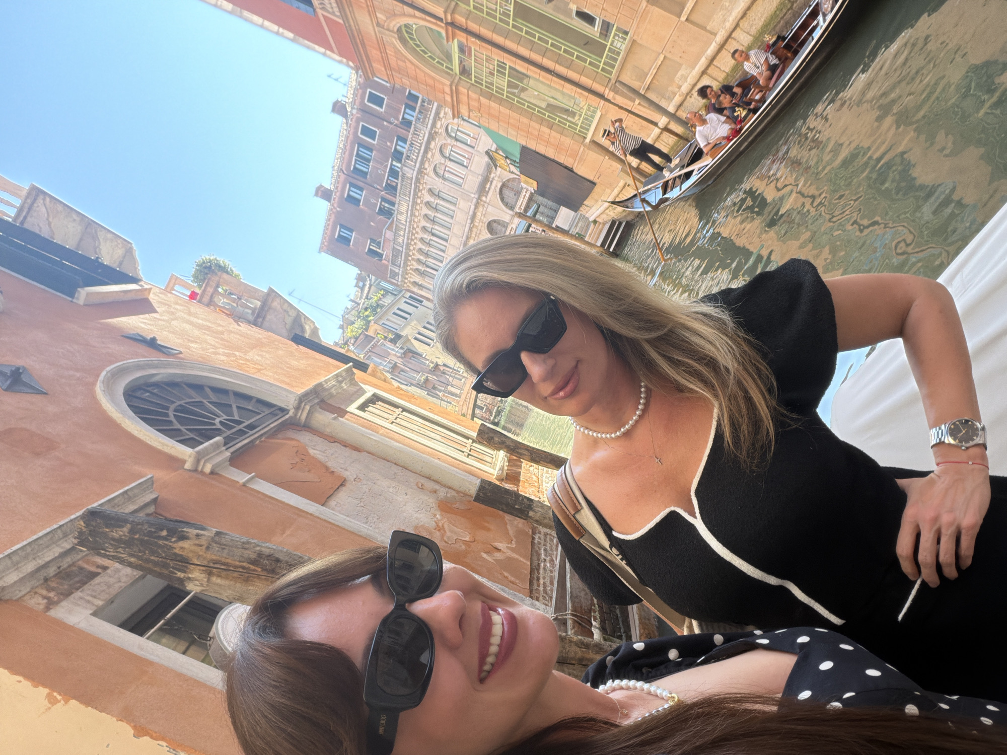 This is a selfie taken by two women in Venice, Italy, in front of a canal. The woman on the left is wearing a black and white polka dot dress and sunglasses. The woman on the right is wearing a black top with white trim, pearls, and sunglasses. They are both smiling. In the background, a gondola with several people is visible on the canal. The buildings in Venice are tall and narrow, with orange and brown facades. The sky is bright blue.