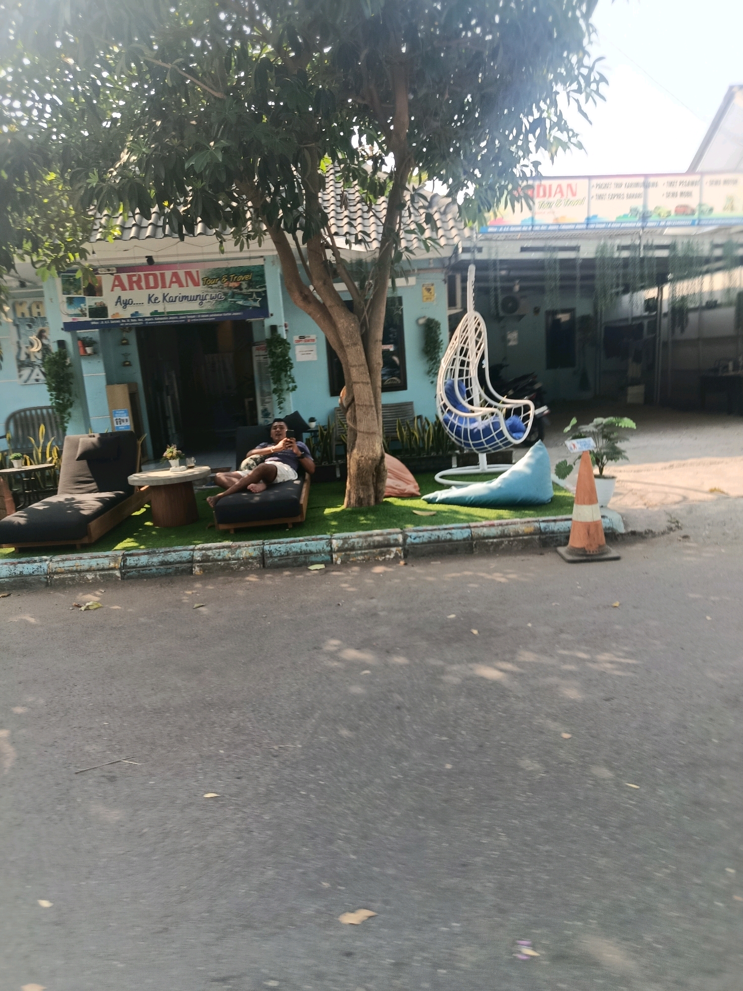 The image captures a scene in front of "ARDIAN Tour & Travel" located in Jepara, Indonesia. In the foreground, a paved road is visible, with fallen leaves scattered across it. To the right, an orange and white traffic cone stands near the curb. Beyond the curb, a small patch of artificial turf hosts a light blue beanbag chair and a white hanging chair with blue cushions. A man is relaxing on a dark grey lounge chair, holding a cigarette in his right hand. He is wearing a dark t-shirt and light-colored shorts. Next to him is another dark grey lounge chair and a small round table with a potted plant. Behind the seating area, the building features a light blue facade with signage for "ARDIAN Tour & Travel" displaying text such as "Ayo... Ke Karimunjawa". Trees with lush green foliage overhang the building and the seating area, casting shadows. The background shows the entrance to the establishment and other structures, with a glimpse of what appears to be a covered parking area or walkway. The overall impression is a relaxed outdoor waiting or relaxation area for a travel agency.