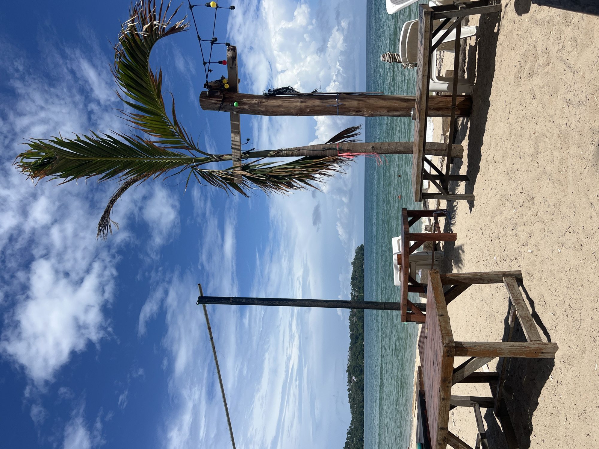 This is a photograph of an outdoor seating area on a beach in Choeng Thale, Thailand. The scene depicts several wooden tables and benches set up on the sand. In the background, the turquoise ocean stretches to the horizon, meeting a bright blue sky dotted with white clouds. A palm tree stands tall to the right of the frame, its fronds extending outwards. String lights with colorful bulbs hang from a wooden post near the palm tree. The beach appears to be a part of a resort or restaurant, with chairs visible in the background. There are no people in the immediate foreground, but two individuals can be seen swimming in the ocean. The overall atmosphere suggests a relaxed, tropical setting.