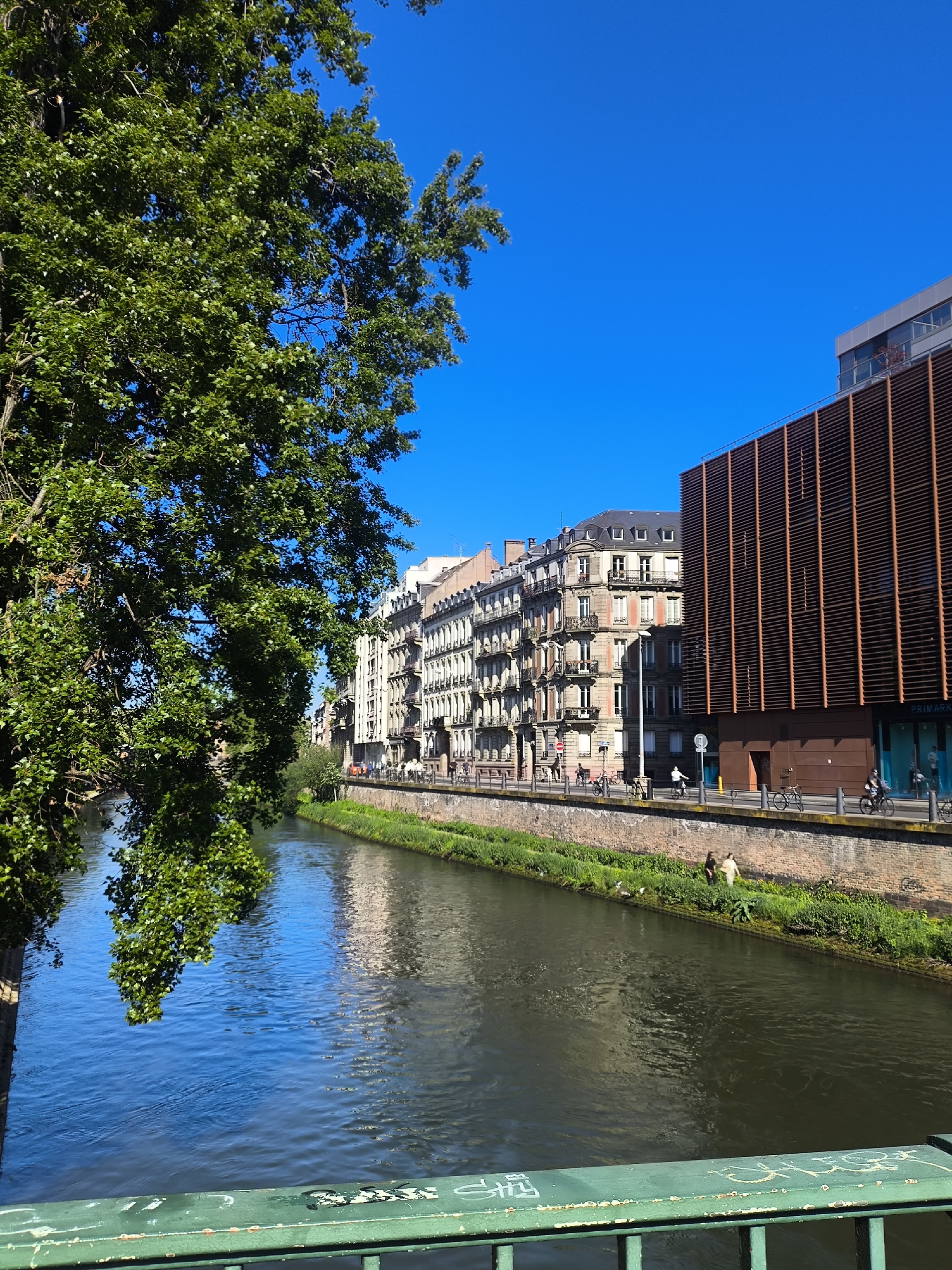 The image shows a canal in Strasbourg, France. On the right bank of the canal, there's a brick-lined path with people walking and cycling. A modern building with vertical wooden slats stands adjacent to older, traditional buildings. Lush green trees are on the left of the frame, partially obscuring the view of the canal. The canal water reflects the buildings and sky. A green metal railing is visible in the foreground. The sky is clear and blue.