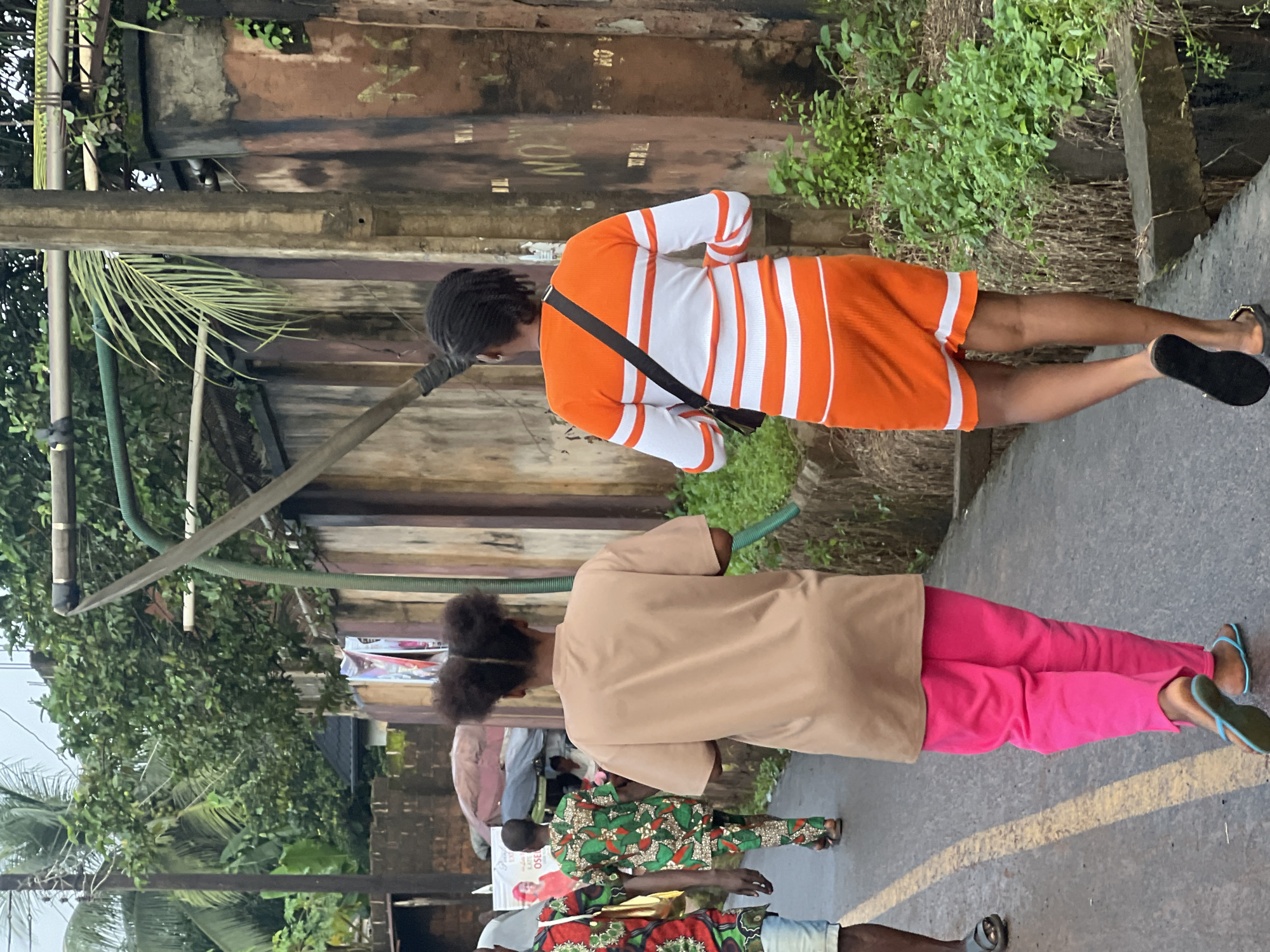 In Nkpor, Nigeria, two women walk away from the camera on a wet asphalt road. The woman on the left wears a tan t-shirt and bright pink trousers, with her hair styled in two buns. The woman on the right is dressed in an orange and white horizontally striped dress and carries a black bag over her shoulder. They are walking past a weathered, multi-colored wall and lush green vegetation. Another man, partially visible on the far left, wears a patterned shirt and denim shorts. In the background, a vendor stall with a red and white umbrella is visible. The scene suggests an overcast day, indicated by the diffused lighting and wet ground.