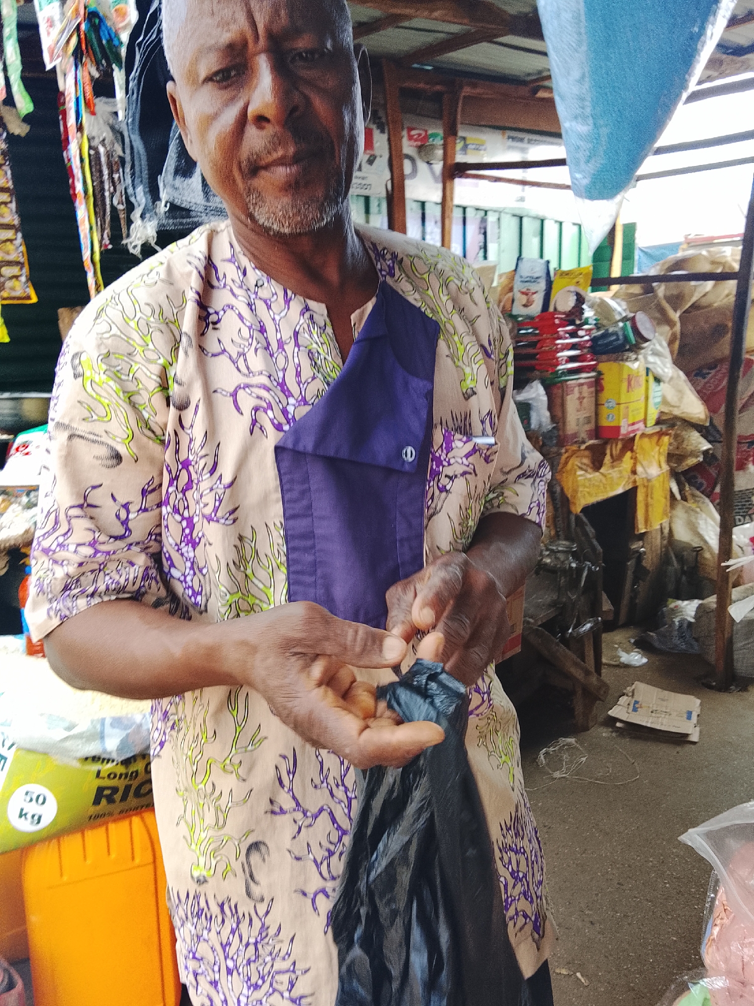 The image depicts a medium shot of an older man in Akwanga, Nigeria, standing in what appears to be a market stall. He has short, graying hair and a beard, and is wearing a light-colored shirt with a purple, coral-like pattern and a dark purple accent panel on the chest. His hands are in the foreground, holding a black plastic bag. Behind him, shelves and piles of goods are visible, including bags of rice labeled "50 kg Long Ric," various colorful packaged items, and what look like sacks of produce. The setting suggests an outdoor or semi-enclosed market environment, with wooden structures and a corrugated metal roof visible in the background. The lighting is natural and appears to be daylight.