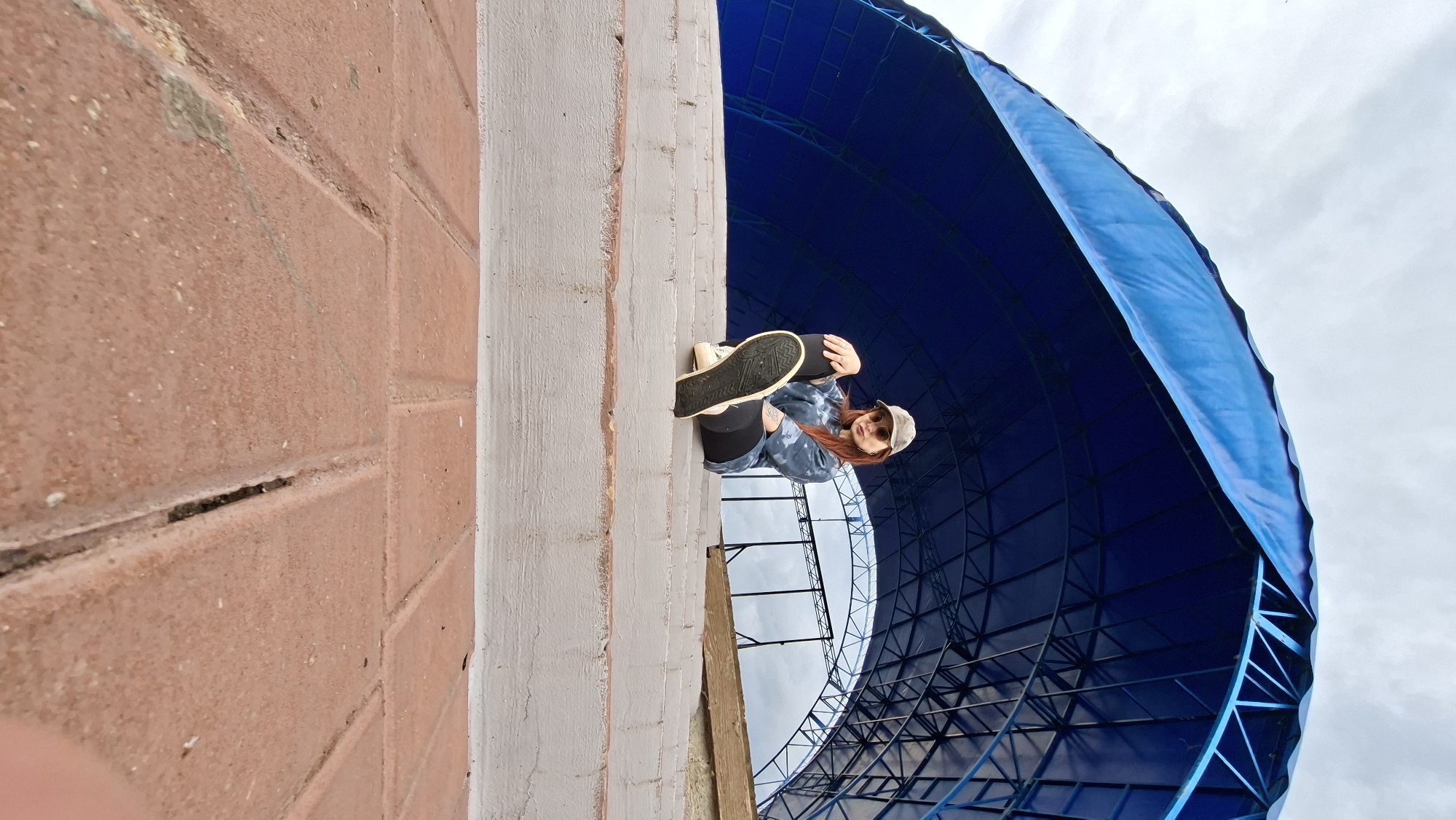 A young woman with reddish-brown hair, wearing sunglasses and a grey baseball cap, sits on a concrete staircase outdoors. She has on a tie-dye sweatshirt and black leggings, with white sneakers. Her right leg is extended forward, with the sole of her shoe visible. Behind her, a large blue arched canopy structure with a metal frame stretches across the background. The scene is lit by daylight under an overcast sky. The foreground shows red brick paving. The location is identified as the Myadzyel City Park, Belarus.