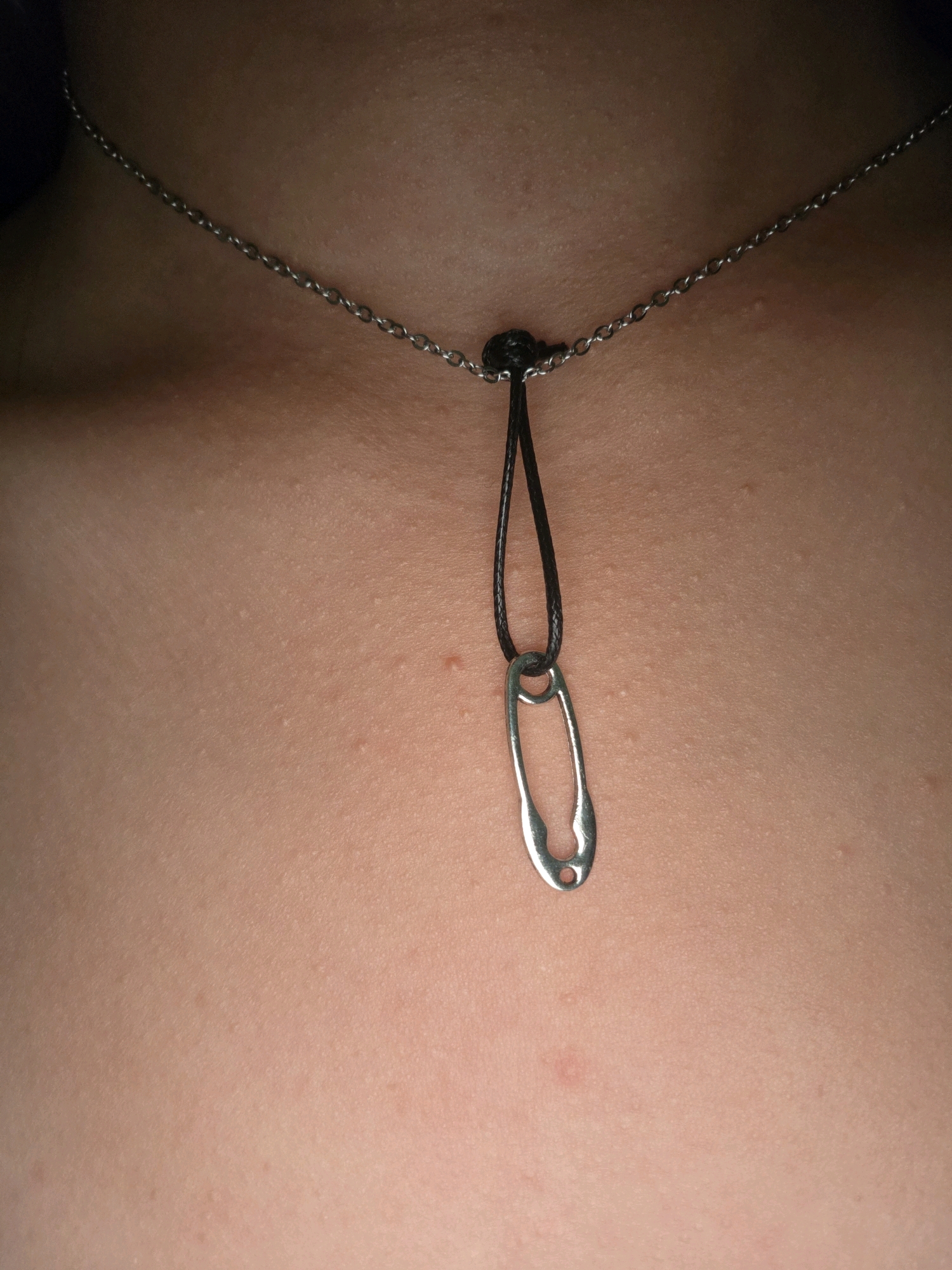 A close-up, eye-level shot focuses on a person's chest, adorned with a simple necklace. The necklace features a delicate silver chain that rests against the skin, with a black cord suspended from it. Dangling from the cord is a silver safety pin, serving as a pendant. The skin appears to have a textured surface with some small, light-colored bumps and a few reddish spots. The lighting suggests an indoor setting, possibly a room with artificial light casting subtle shadows and highlights on the skin and jewelry. The background is out of focus, with the primary subject being the necklace and the skin. The image is cropped tightly, excluding any facial features or the rest of the person's body. There is no visible indication of the city of Phan Thiết, Vietnam, or any urban environmental elements.