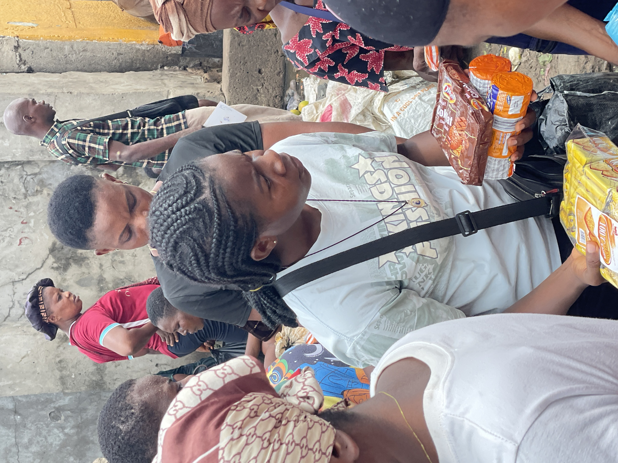 The image depicts a crowded gathering of people, primarily adults, in what appears to be a public, possibly semi-outdoor, waiting area in Onitsha, Nigeria. The ground is concrete, showing some wear and a faded yellow line, indicating an utilitarian setting like a market, bus station, or transit point.
A woman with braided hair wearing a light blue t-shirt is prominent in the foreground, looking upwards with a neutral expression. Text partially visible on her shirt includes "Separa" and "KINGS SHION". She has a black strap from a shoulder bag across her chest. Her hand holds several packaged snack items: a brown packet of "Coklat" (chocolate) wafers or biscuits, and two orange-capped cylindrical containers. Nearby, a large yellow package, possibly of biscuits, displays "100 C".
Surrounding her are numerous other individuals. Behind her, a person in a black t-shirt is visible. To the left, a woman in a red t-shirt with a dark head covering is present. Further back, a bald man in a green and brown plaid shirt stands or sits. Other people are seen with various head coverings, including one with a brown and white patterned fabric, and another in a white t-shirt in the bottom right. A white sack bag with text "IN A COOL POY PE" is visible on the ground, suggesting luggage.
The scene conveys a sense of waiting or resting, with the packaged food items hinting at travel or a prolonged stay. The natural lighting indicates it is daytime. Emotions generally appear subdued or pensive among the individuals.