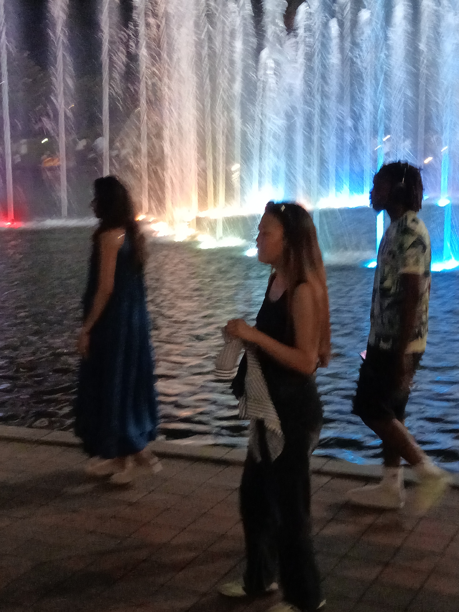 The image shows a night scene with a fountain in the background. The fountain has water jets illuminated with colorful lights, including red, orange, and blue. In the foreground, three individuals are visible. On the left, a person in a dark blue dress walks away from the viewer. In the center, a person with long brown hair wears a black top and dark pants, holding a striped cloth. To the right, a person with dark, curly hair wearing a patterned short-sleeved shirt and dark shorts stands facing away from the viewer. The ground appears to be paved. This scene is likely located at the KLCC Park in Kuala Lumpur, Malaysia, known for its Symphony Lake and fountain displays.