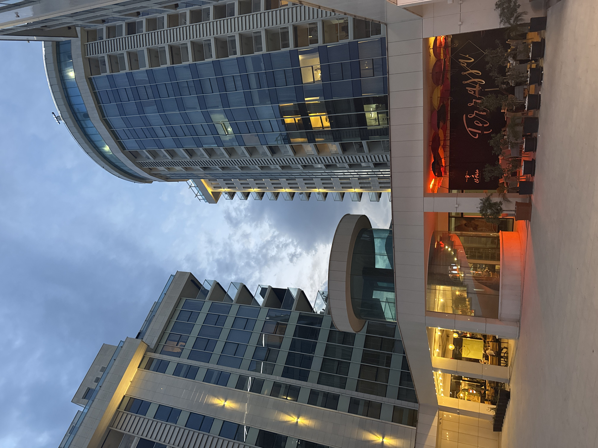 This image captures a modern urban scene in Budva, Montenegro, likely during late afternoon or early evening. The sky is partially cloudy with shades of blue and gray, hinting at dusk.
Two prominent, contemporary high-rise buildings dominate the frame. On the left, a multi-story building features a glass facade and stepped balconies. On the right, a taller, distinctively curved building boasts a blue-tinted glass exterior and numerous balconies. Reflections are visible on its glassy surface, and artificial lights illuminate several windows, indicating occupancy. A unique cylindrical architectural element protrudes from this building.
At the base of the rightmost building, an inviting outdoor dining or lounge area is visible. It features tables, chairs, and potted plants, creating a pleasant ambiance. A large, dark sign is prominently displayed, bearing the stylish script "Terrassa" in a light color, with "El Faro" written below it. Red and orange lighting adds warmth to this area, and a pattern of colorful umbrellas suggests a shaded or decorative overhead element.
While no specific individuals are clearly discernible, the illuminated windows and the set-up of the outdoor area imply ongoing activities such as dining, socializing, or residential living. The modern architecture and the presence of a "Terrassa" (terrace restaurant) align with the context of a resort city like Budva.