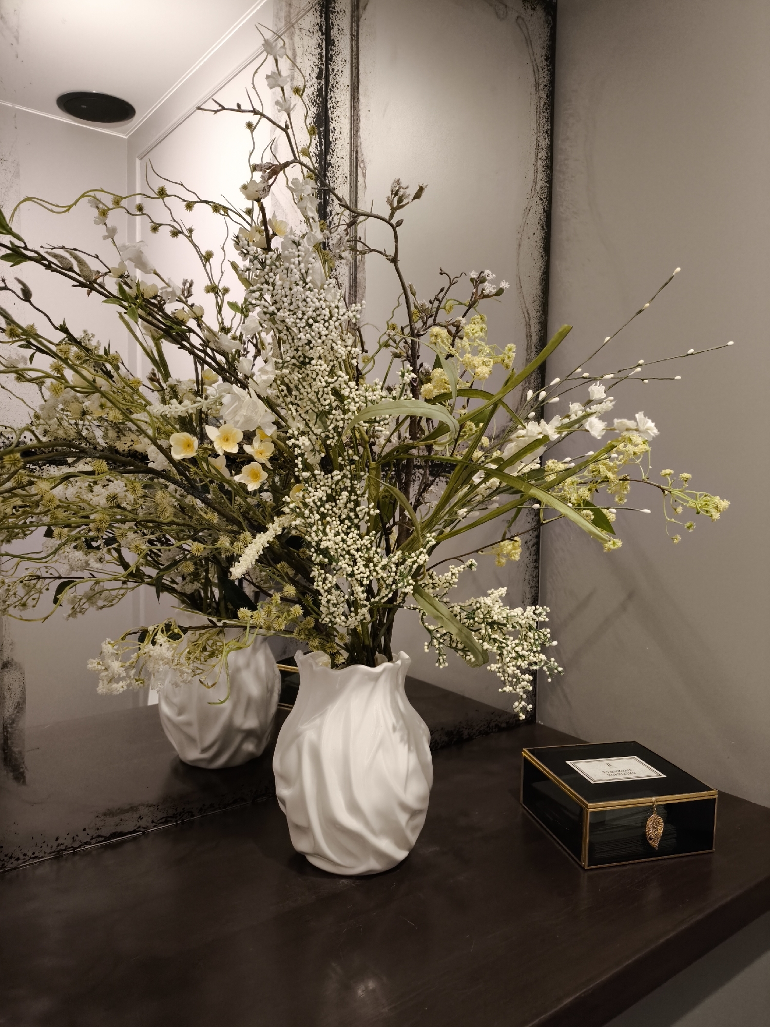 A still life presents a bouquet of flowers arranged in a white vase. The flowers are a mixture of greenery, small white blossoms, and yellow-centered white flowers, filling the vase to overflowing. A reflection of the bouquet and vase can be seen in the mirror placed behind it. To the right of the vase sits a small black and gold box. The scene is set on a dark surface, perhaps a table or counter. The wall is a neutral grey color. The location is not Smolensk, Russia.