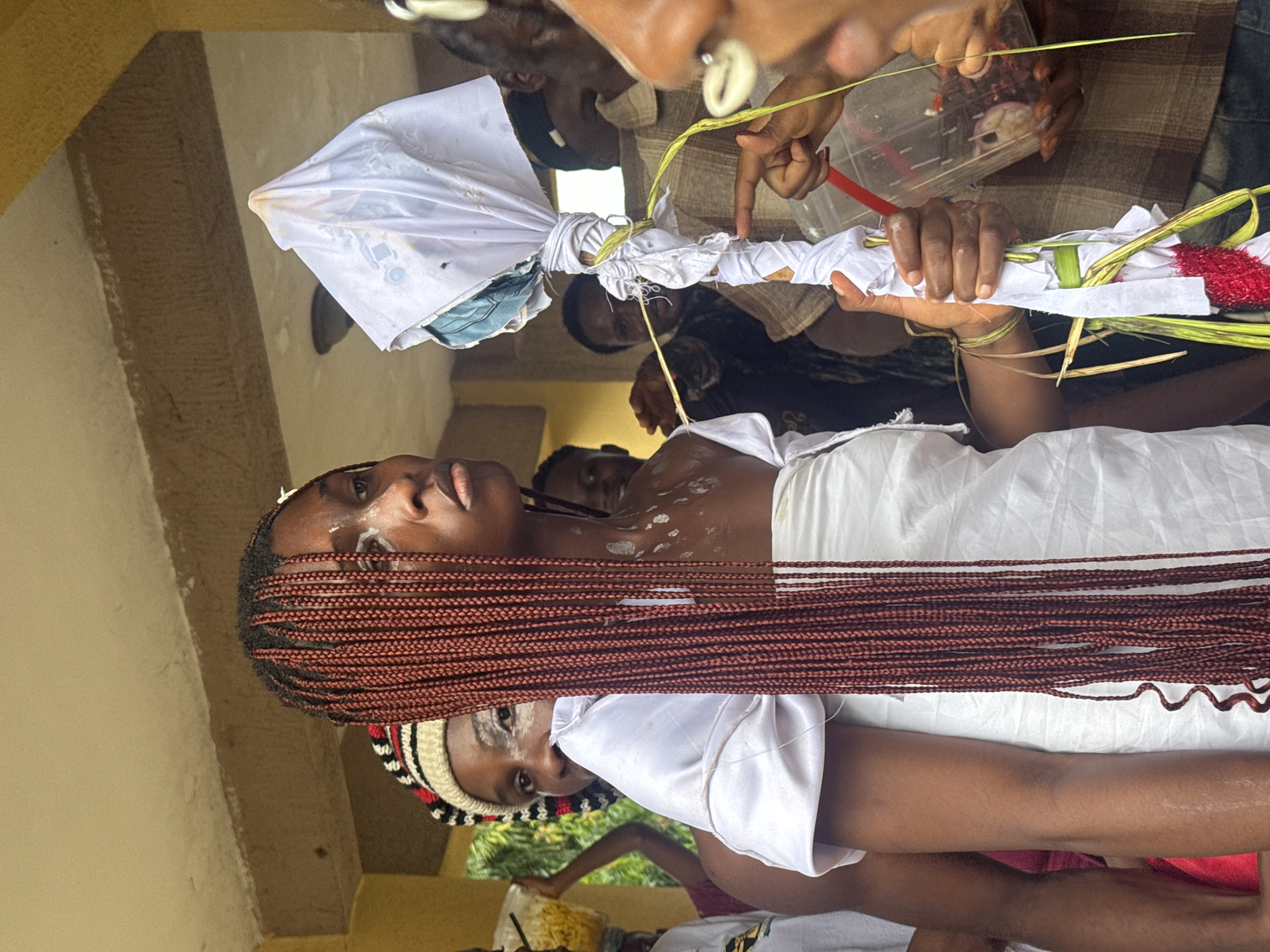 In Ihiagwa, Nigeria, a group of people are gathered outdoors under a covered walkway. The central focus is a young woman with long, reddish-brown braids, wearing a white garment. White markings are visible on her face and chest. She is holding a staff-like object wrapped in white cloth and decorated with green leaves and red fabric at its base. Another individual's hand, adorned with white shells, is near the staff, seemingly pointing to it. Behind the central figure, other individuals are partially visible, some looking towards the front. In the background, a person is seen carrying a large bag of yellow items. The environment appears to be a communal space with a concrete floor and overhead structure.