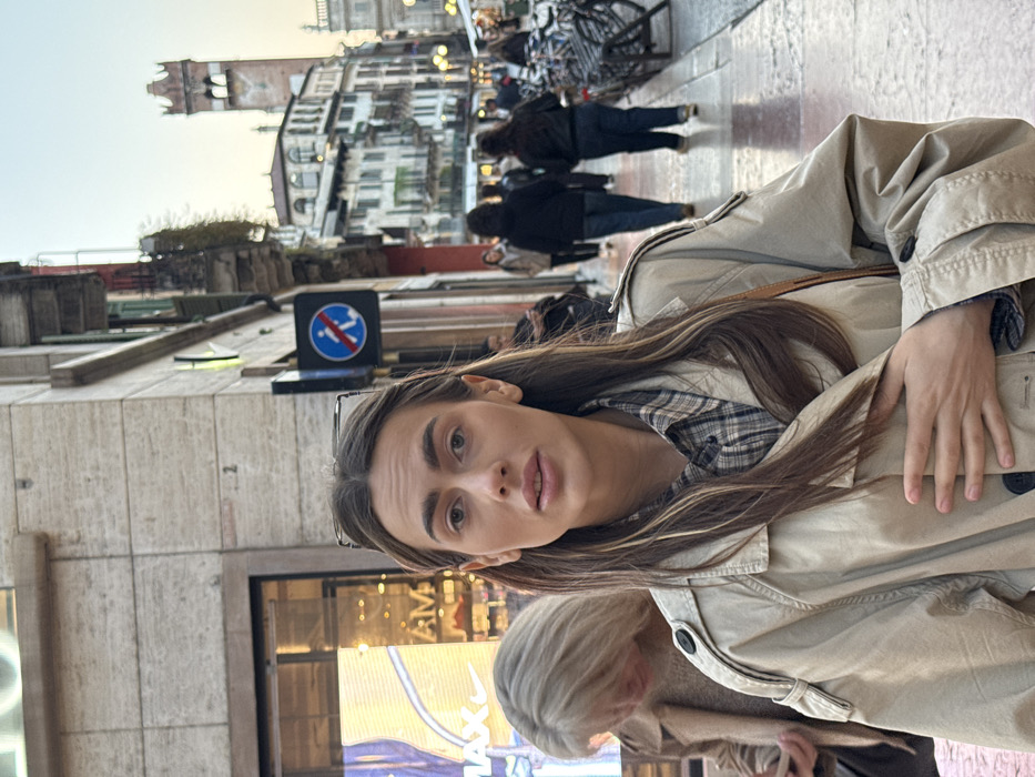 The scene depicts a street in Verona, Italy, with the Lamberti Tower prominently visible in the background. In the foreground, a young adult female with long, dark brown hair and clear-rimmed glasses resting on her head looks directly at the camera. She is wearing a light beige trench coat over a plaid shirt, with her right hand placed flat on her chest. To her immediate left, another female with lighter-colored hair is partially visible, facing downwards. The building on the left features large glass storefronts, one displaying "Nike" and "Air Max" branding, indicating a retail establishment. To the right of the main subject, a blue circular sign with a white diagonal bar and a figure, indicating a pedestrian restriction, is mounted on a pole. Further down the street, multiple blurred figures of pedestrians are observed walking away from the camera, and a cluster of bicycle racks with bicycles is present on the right sidewalk. Distant buildings with traditional Italian architecture line the thoroughfare, culminating in the red brick Lamberti Tower with its crenellated top, standing against a late afternoon or early evening sky. Artificial lights illuminate the storefronts and some distant buildings.