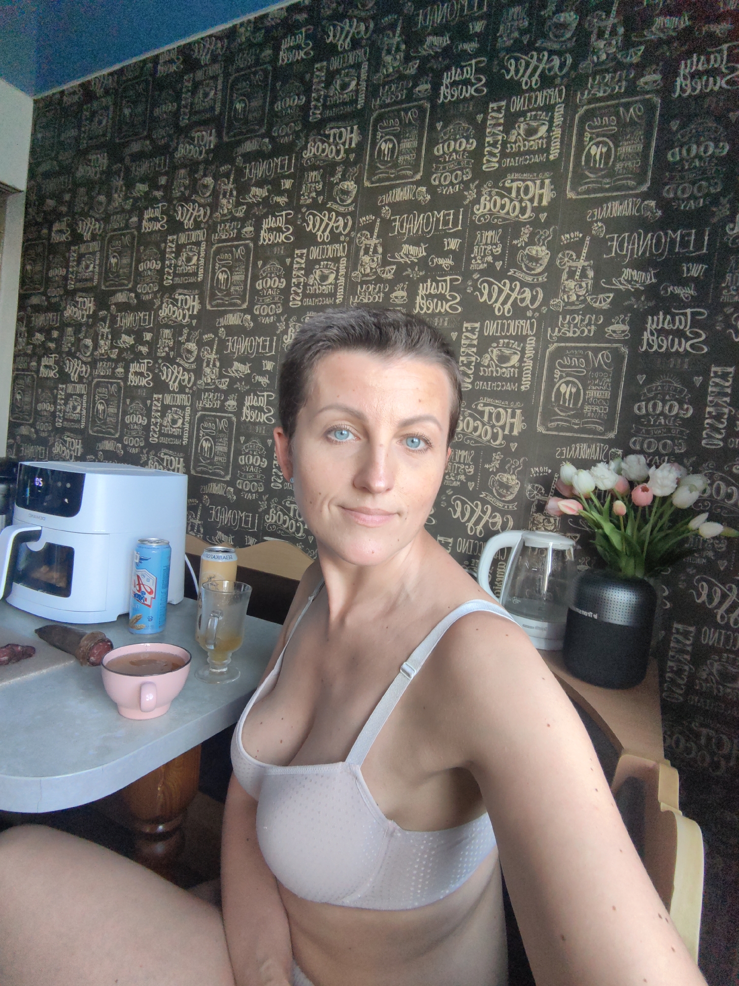 In this media file, a woman is featured in a domestic setting, likely a kitchen or dining area. She is seated at a table, facing the camera with a neutral expression. Her short, dark hair frames a face with striking blue eyes. She is wearing a light-colored bra.
On the table in front of her, several objects are visible. To her left, a white air fryer with a digital display showing "05" is prominent. Next to it are two cans, one predominantly blue and white with some text, and another taller, lighter-colored can. A glass with a yellow liquid and a handle, resembling a mug or a traditional beer glass, stands beside the cans. A pink ceramic bowl filled with a brown liquid, possibly a beverage, is also on the table, alongside a slice of what appears to be salami or a similar cured meat on a cutting board.
To the woman's right, a glass electric kettle and a vase filled with a bouquet of tulips—white, pink, and red—are positioned. The background is covered with patterned wallpaper featuring a repeating design of coffee-related motifs, text, and illustrations, giving a cafe-like ambiance.
The lighting suggests it might be daytime, though no specific time is indicated. The overall impression is a casual, everyday moment captured at home. The woman's posture and gaze convey a sense of self-awareness rather than an overt emotional state. Visible text includes branding on the air fryer and labels on the cans, as well as repeated text and graphics on the wallpaper. The location context of Fanipal, Belarus, provides a geographical reference for this scene.